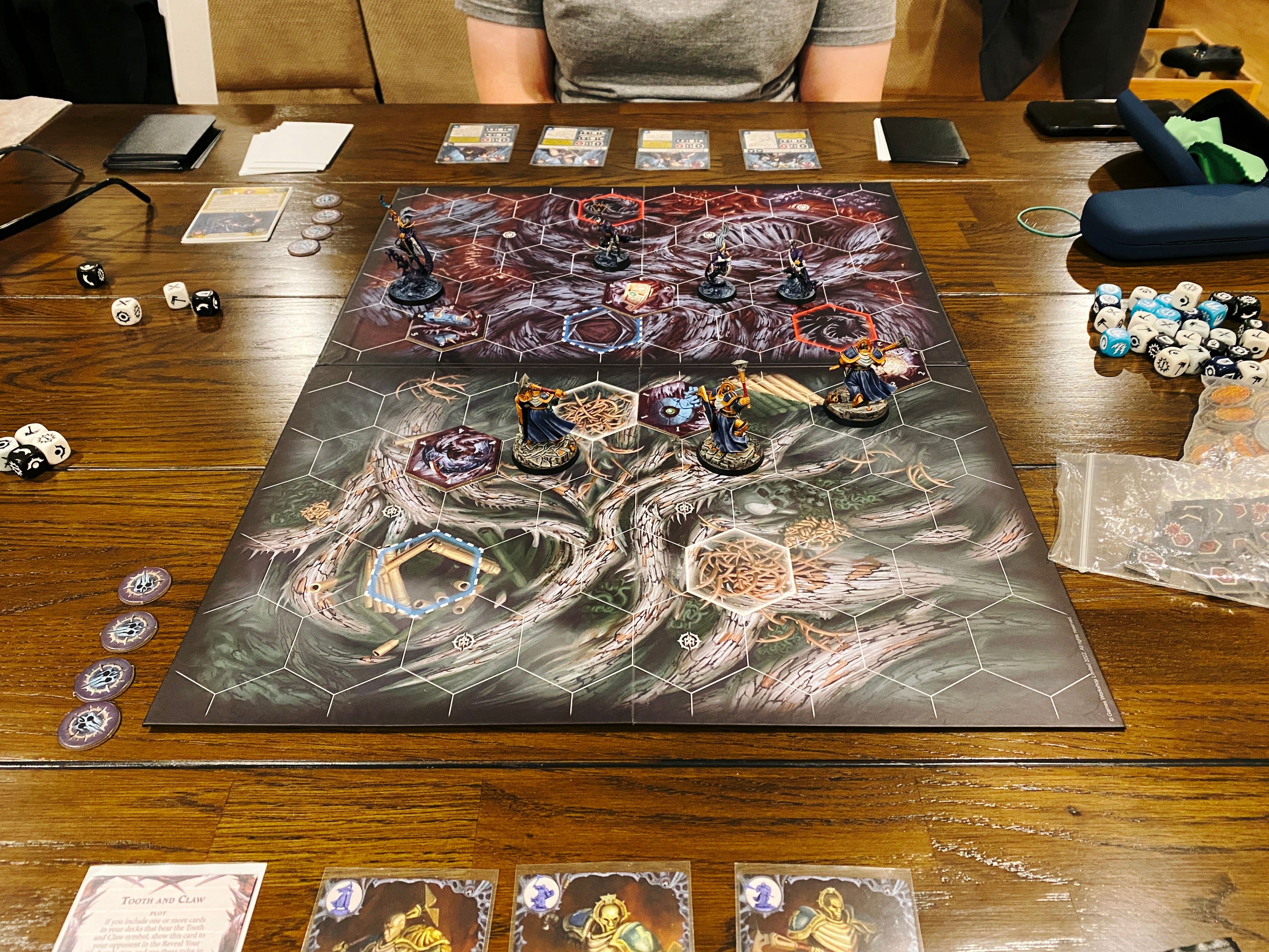 A photo of the game board of a Warhammer Underworlds game. The artwork on it looks like a bunch of gnarled roots and it's covered in hexagons that indicate distances. Closest to the camera are Ironsoul's Condemnors, three heavily-armoured warriors in deep blue and gold armour wielding maces and shields. Facing them are the Shadeborn, four lithe elves in very dynamic poses with very pale skin and wearing deep purple clothing.