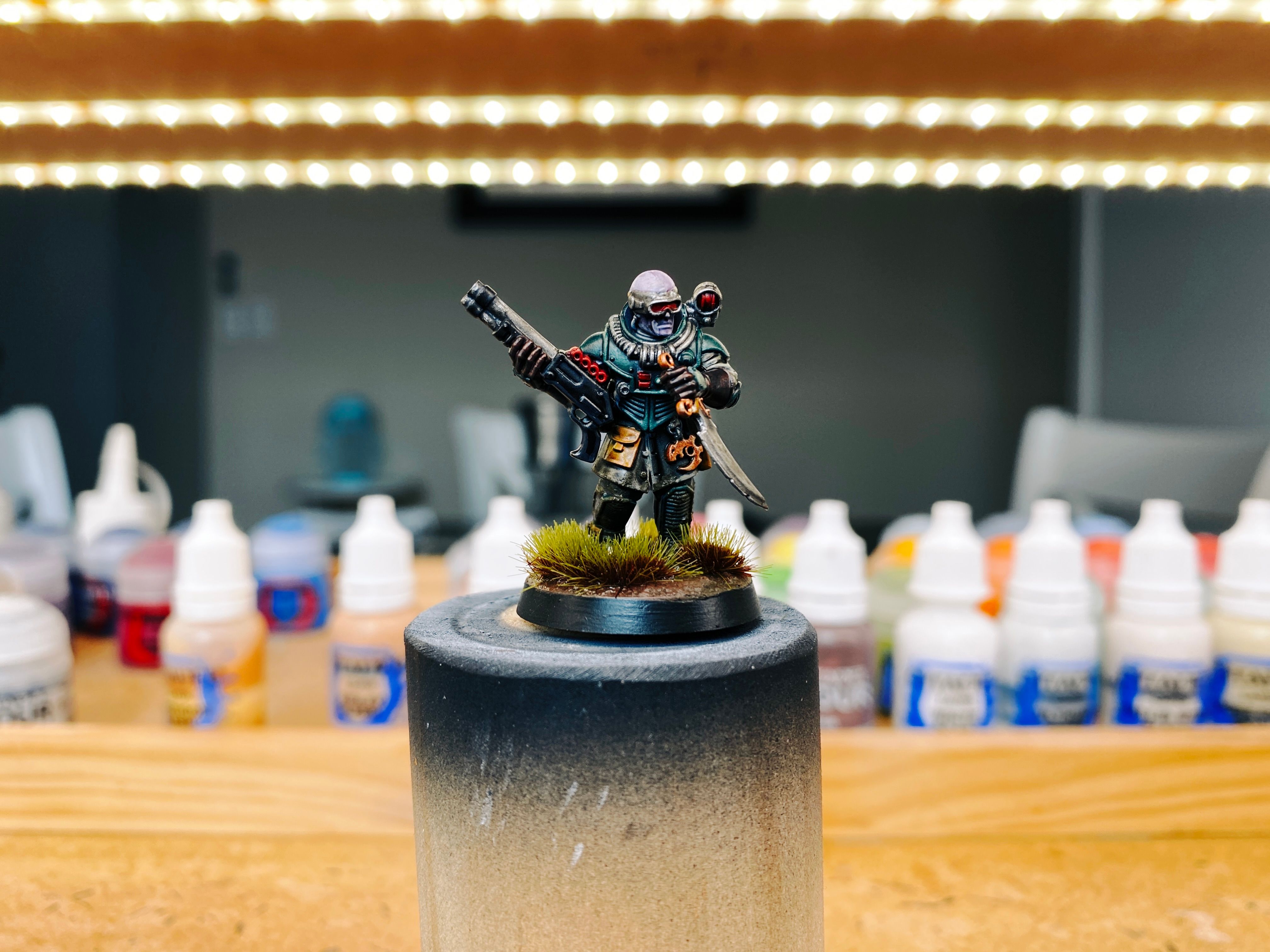 A photo of a Warhammer 40,000 miniature, he's wearing dark green armour and is holding a shotgun in one hand and has a big knife in the other, with the pose implying he's about to use it. He's wearing industrial-looking goggles and looks mostly human except his skin is a very light purple and his skull is just... slightly too big. The base of the miniature looks like it's completely muddy and there's lots of dark green and brown grasses growing, and the mud is splattered up his legs.