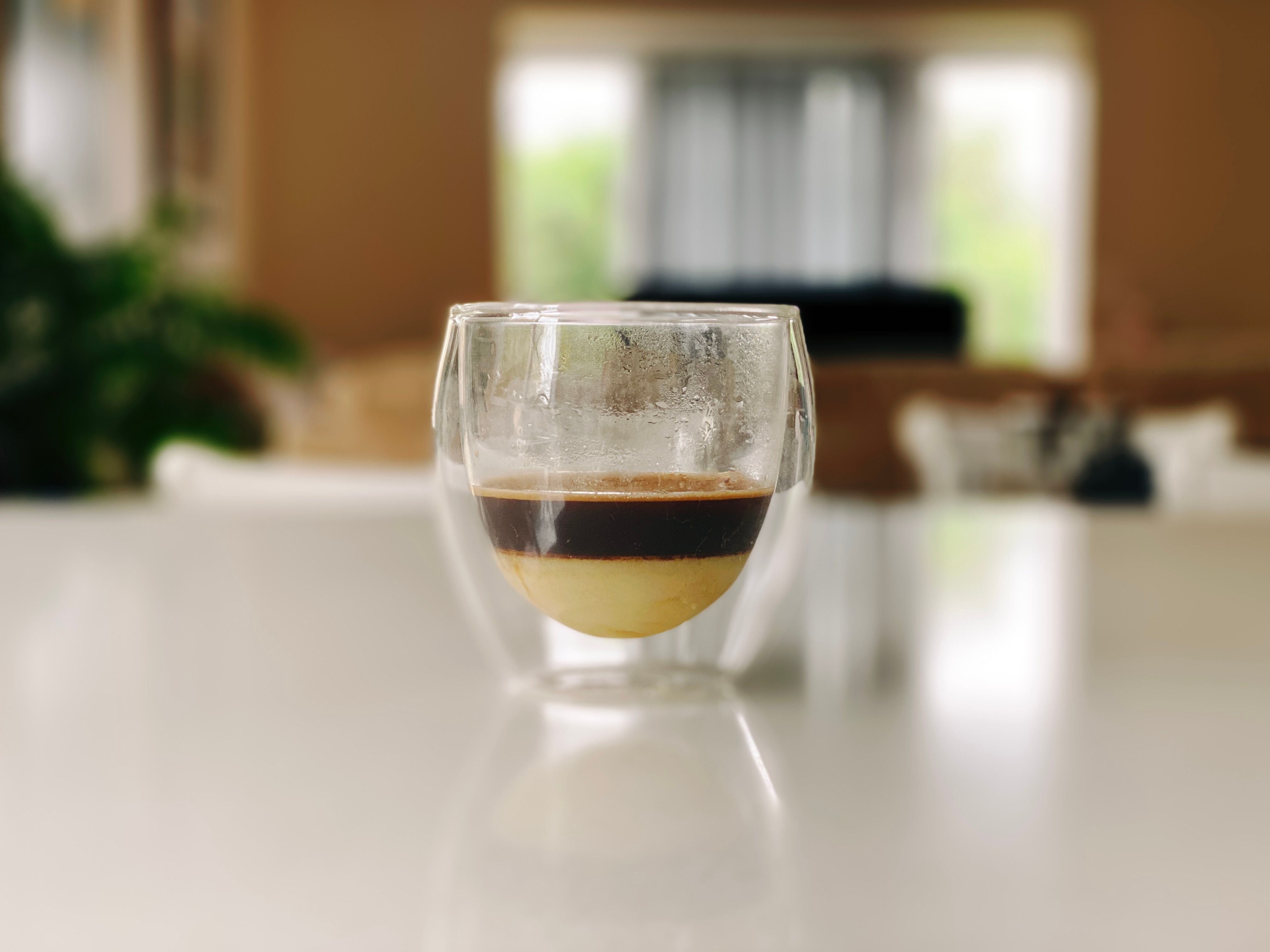A photo of a fancy double-walled round espresso glass about half-way full. The bottom half is a layer of sweetened condensed milk, and the top half is espresso.