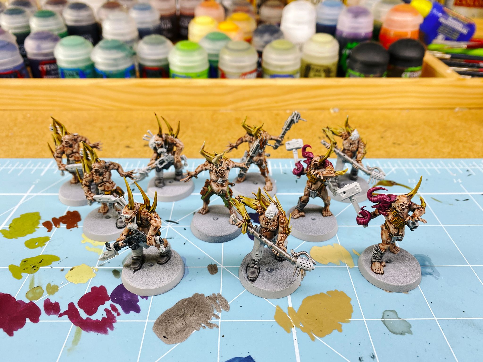 A photo of ten half-painted Nurgle poxwalkers. They're gross distended zombie-looking things with big bone spikes growing out of them, some have pink tentacles, there's lots of pustules all over their bodies, and they're brandishing all sorts of makeshift pieces of industrial equipment as weapons.