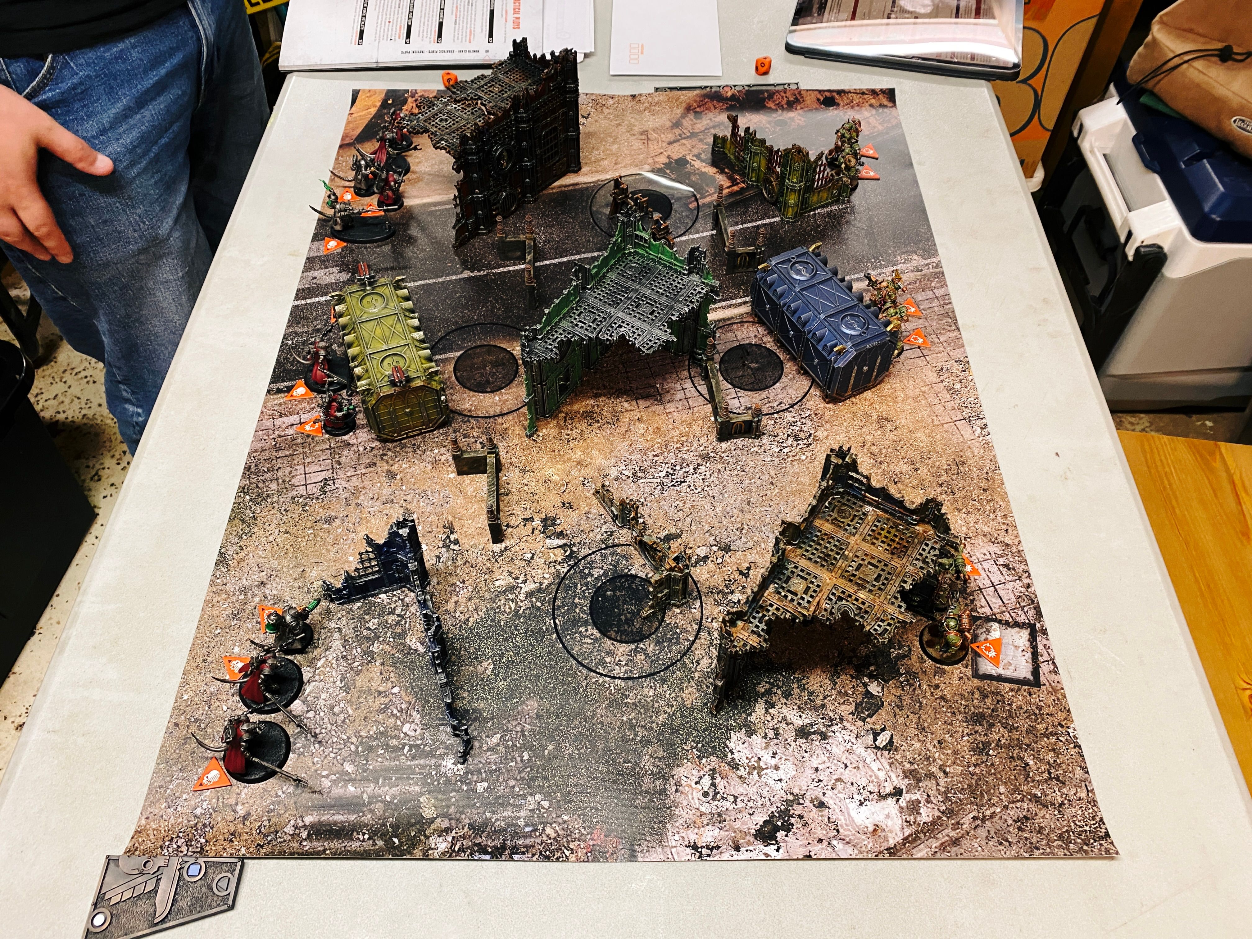 A photo taken from the side of the board of a game of Warhammer 40,000: Kill Team. The terrain is all ruined industrial-slash-gothic buildings, on the left are my Death Guard which are hulking super-humans in a somewhat sickly-green power armour and lots of tentacles and pustules and general grossness, and on the right are the Adeptus Mechanicus who believe in the purity of the machine and replacing as much as possible of their biological components with machine parts and such mostly look like robots with cool cloaks on.

(The AdMech were actually custom 3D-printed models that combined the Adeptus Mechanicus with the Skaven rat-men from the Warhammer fantasy universe, and were really very well done.)