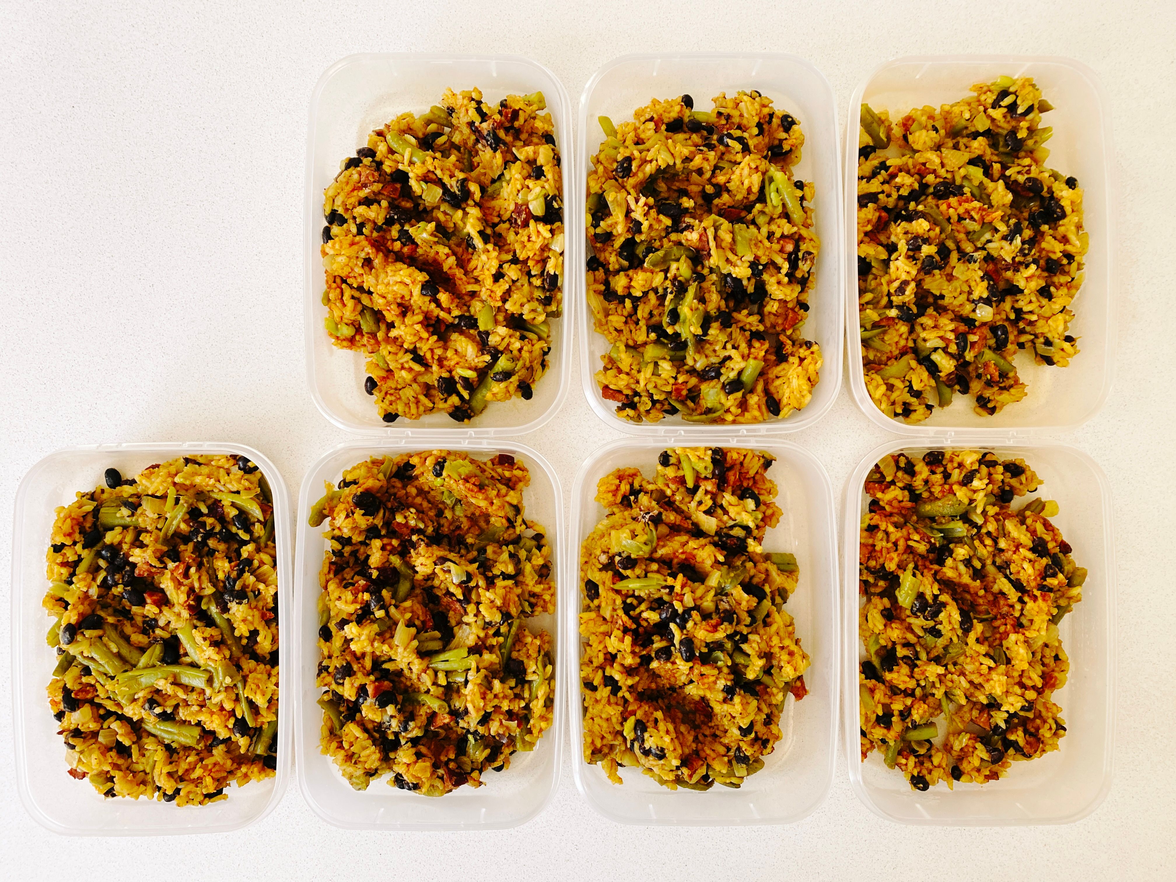 A photo of seven individual tupperware containers with yellow rice in them. There's black beans, green (string) beans, chorizo, and onion in it.