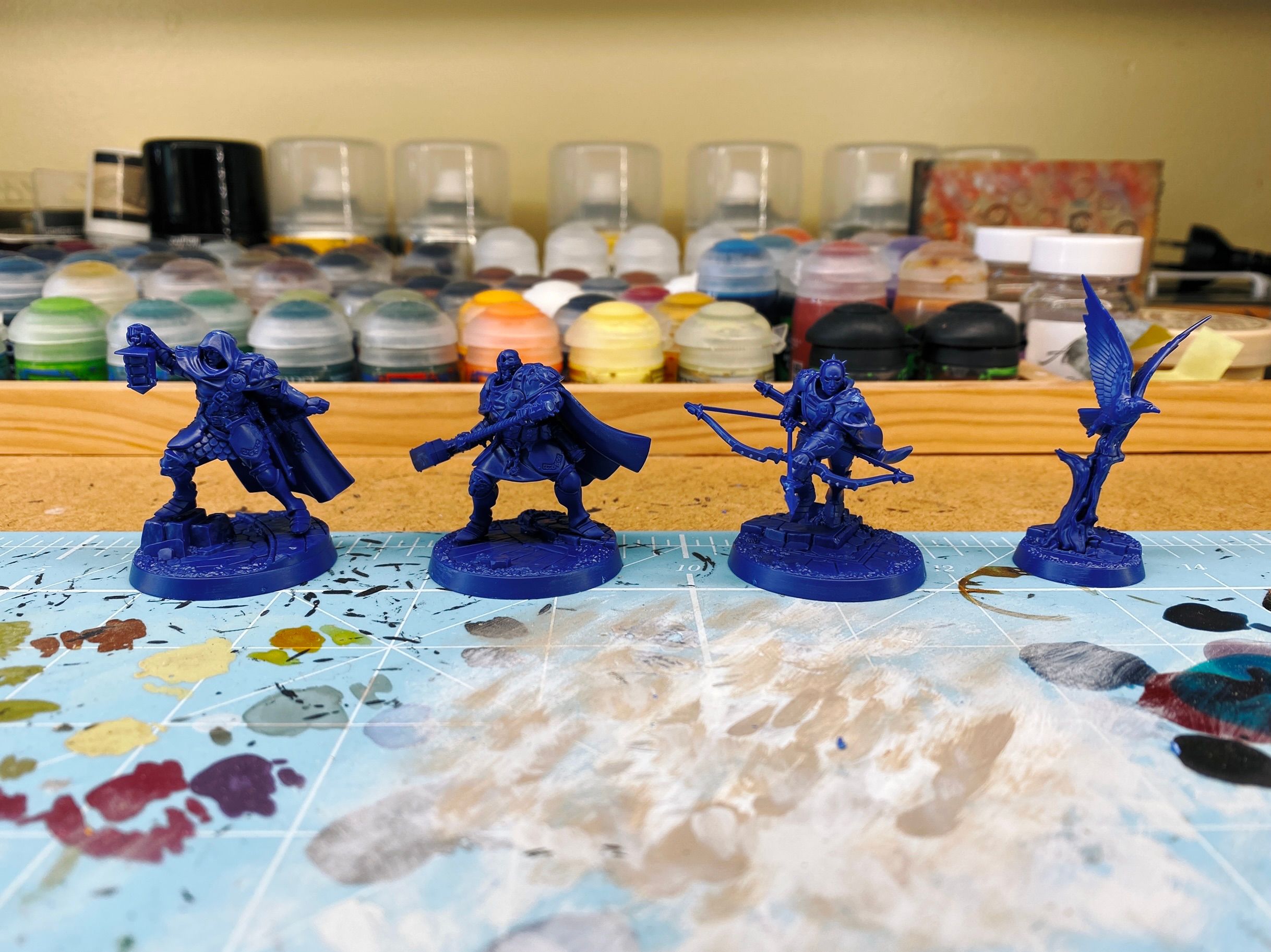 A photo of four blue plastic miniatures. Three are heavily armoured warriors — one with a cloak and holding up a lamp, the second holding a huge two-handed warhammer like he's about to swing it, and the last with bow and arrow in mid-run — and the last is a bird taking flight from a tree.