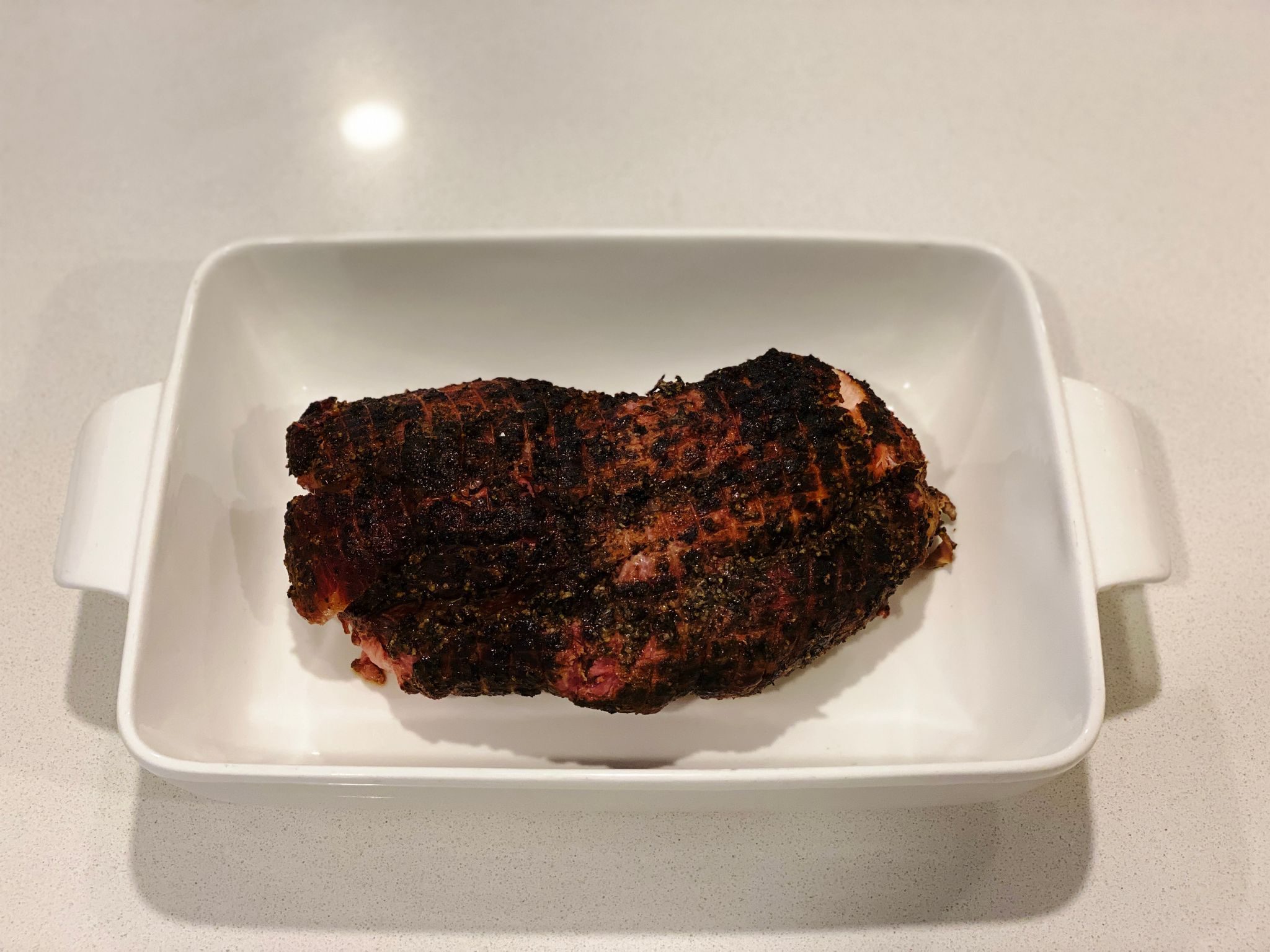 A photo of a fully-cooked and smoked pork collar butt sitting in a white ceramic dish. It's got a fantastic dark colour to the outside.