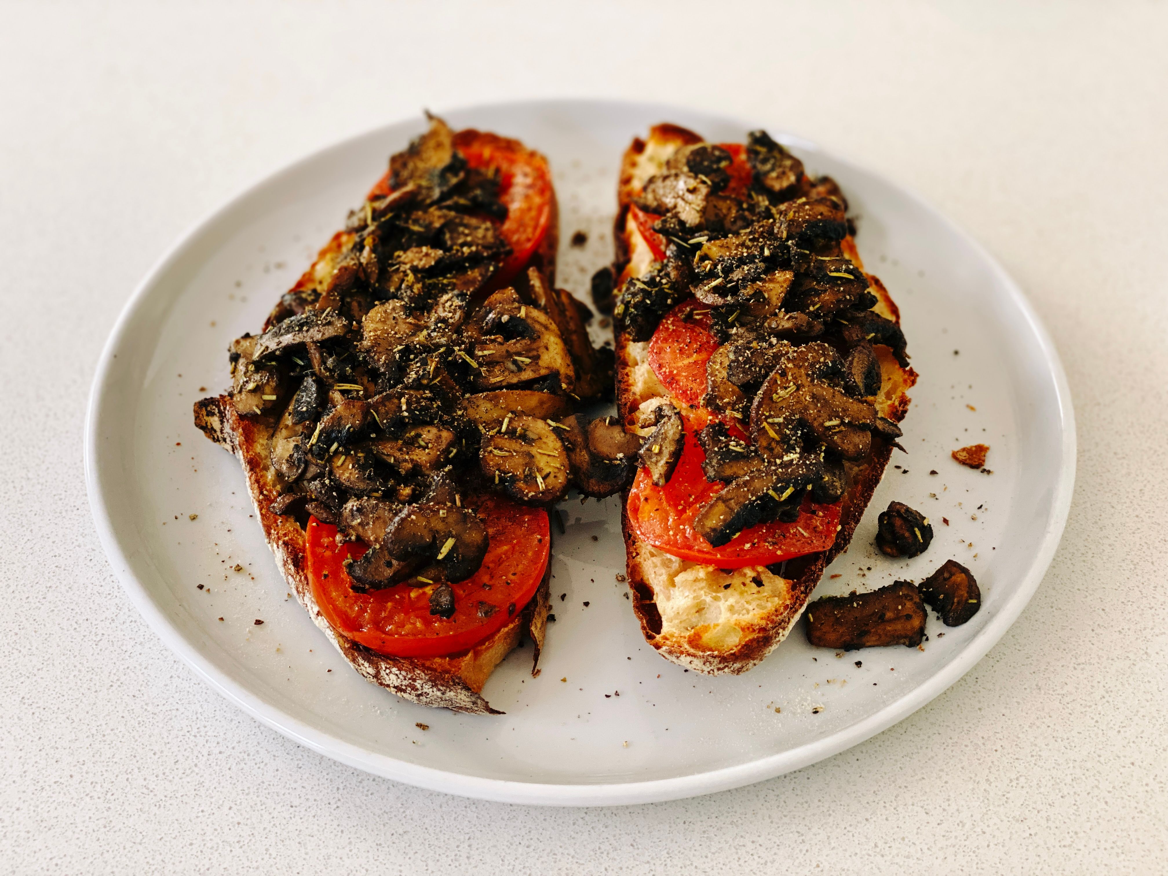 A photo of two slices of the aforementioned bread with slices of cooked tomato on top, and sliced mushrooms cooked in sage, rosemary, and garlic on top of that, all sitting on a white dinner plate.