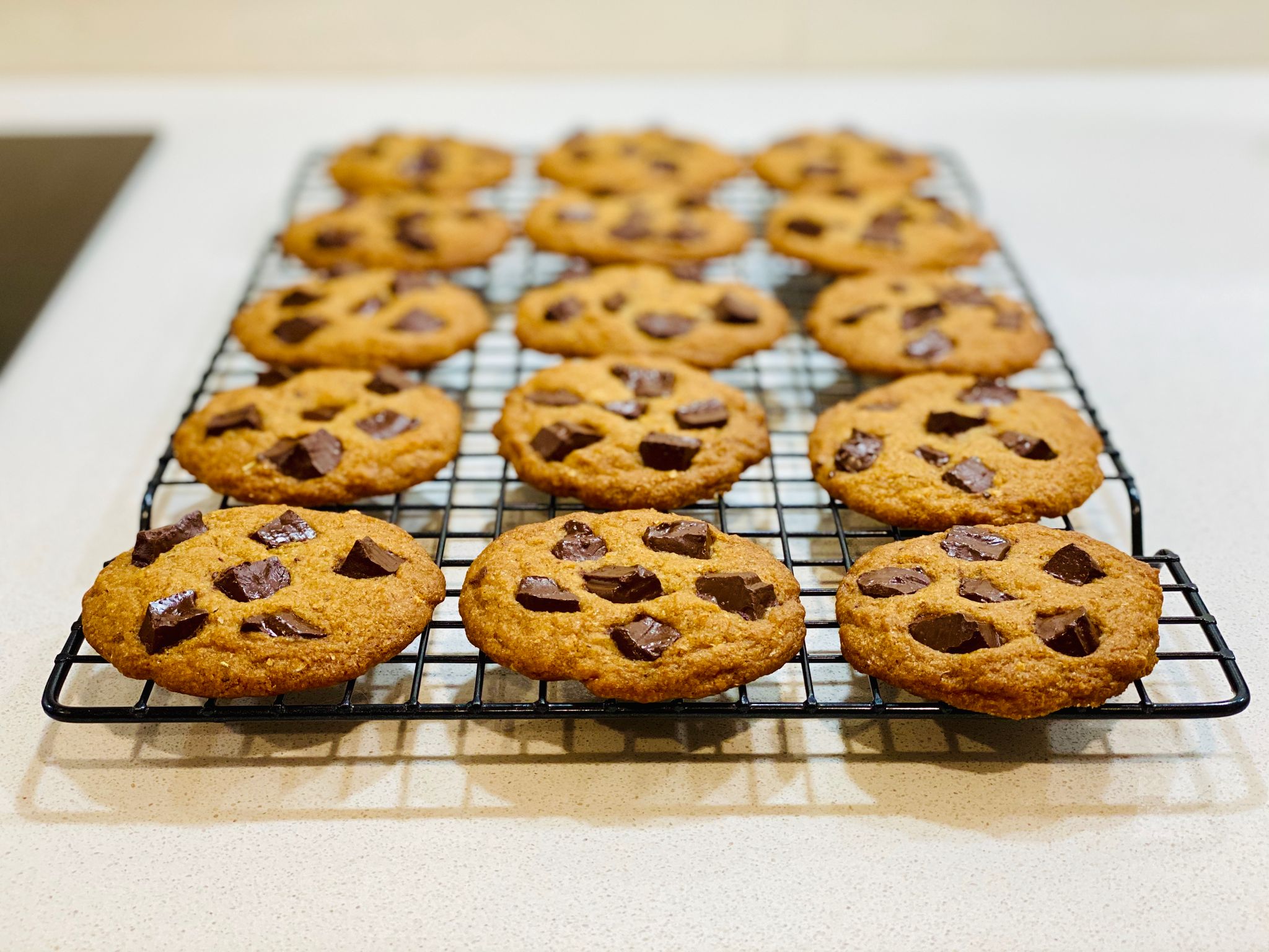 A photo of a batch of cookies sitting on a cooling rack. They're round and very flat, with five chunks of dark chocolate on top of each one.