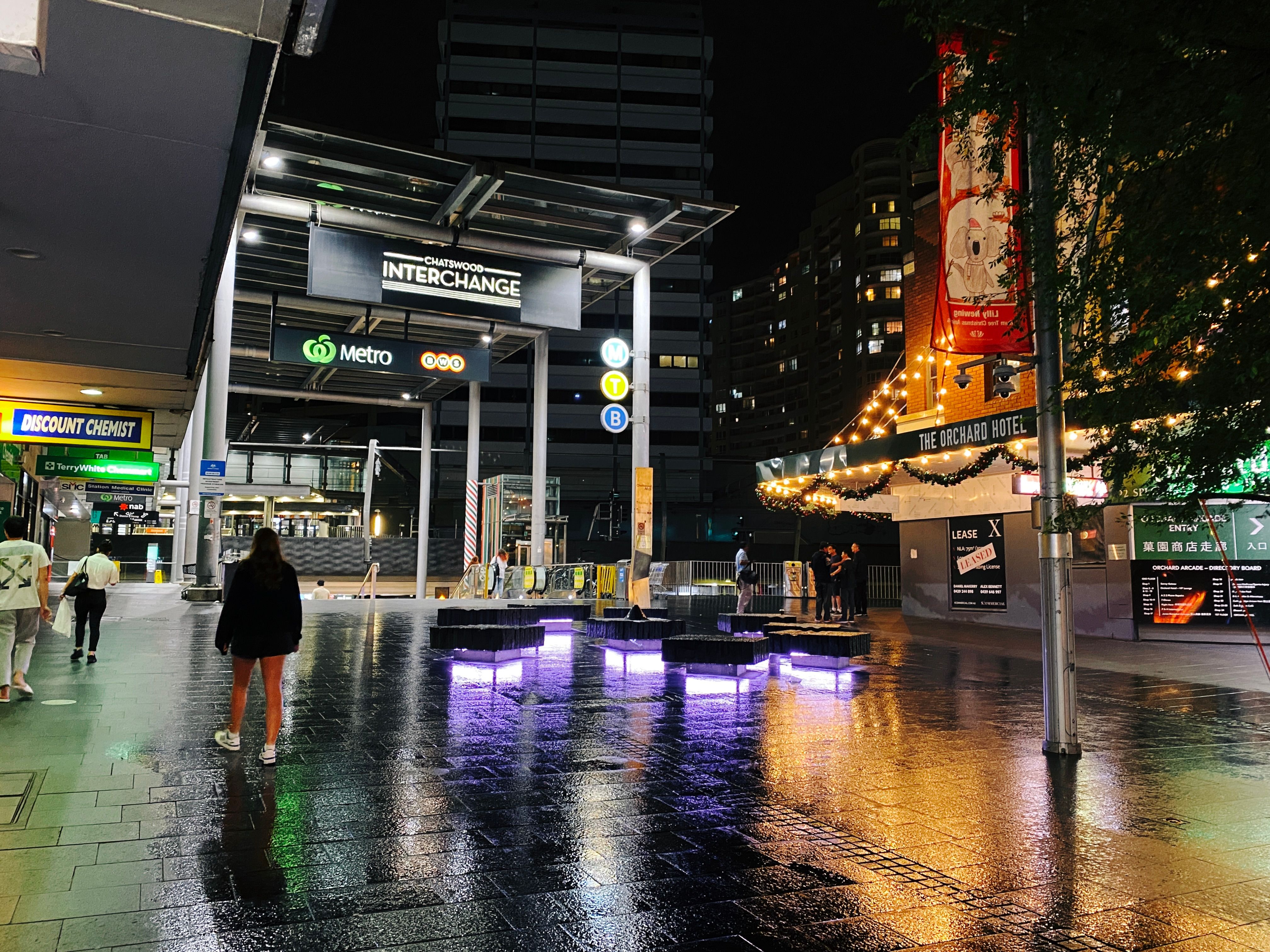 A night-time photo looking at the big entrance arch to Chatswood Interchange where the train station is. It's been raining and there's lots of wonderful reflections of lights on the ground.