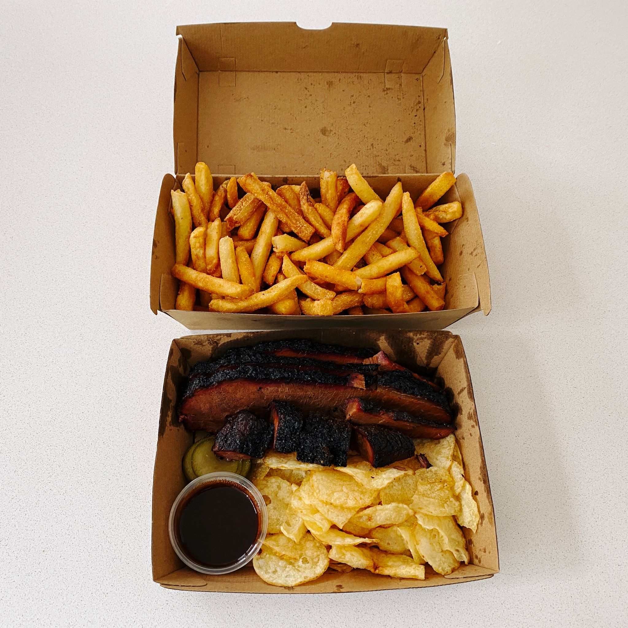 A photo of two cardboard takeaway boxes, one has hot chips, the other has slices of beef brisket, some potato chips, pickles, and a container of barbecue sauce in it.