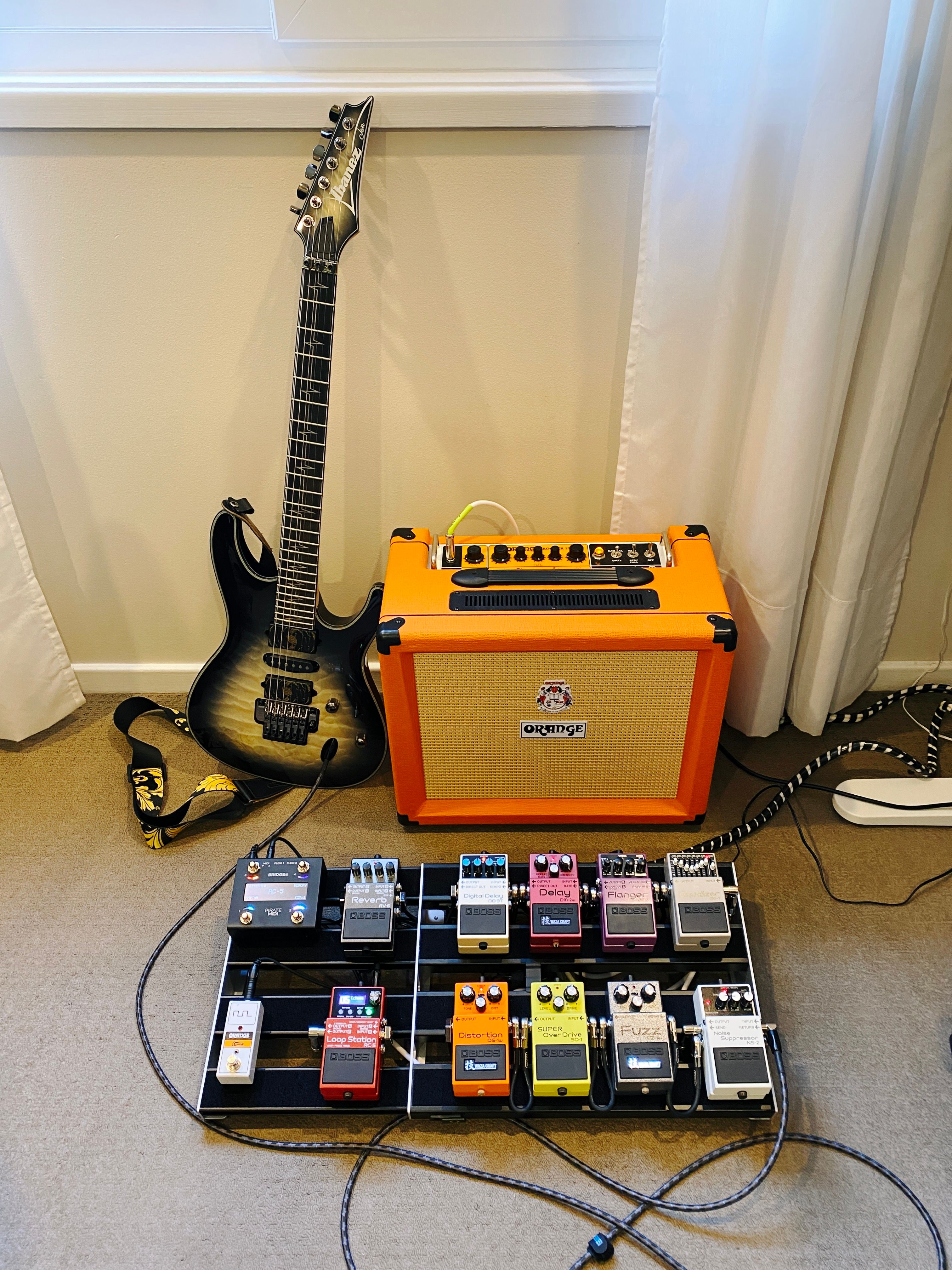 A photo of an Ibanez JIVA10 guitar sitting against a wall next to an Orange amplifier, with a pedal board in front of them with 10 Boss compact pedals on it, plus a MIDI controller pedal and little footswitch pedal for switching the amp between the clean and dirty channels.