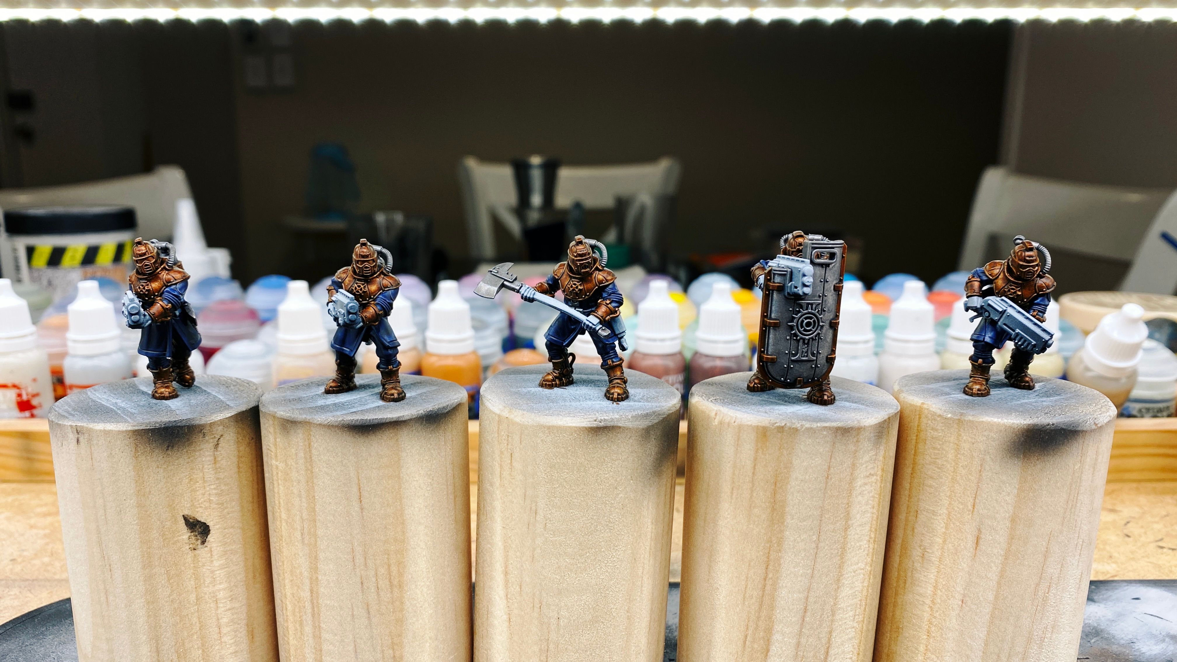 A photo of partially-painted five armoured human warrior miniatures. Their armour is a deep brass colour, they're wearing big mag-boots of the same colour, and their clothes are a rich dark blue. The helmets are reminiscent of diving bells except with a cool-looking visors instead of a round viewing port. Three of them have big short guns, one is wielding a huge axe, and the other has a massive shield that's taller than he is, with a viewing port and a slot for his gun to sit in so he can see and shoot while remaining behind the shield.