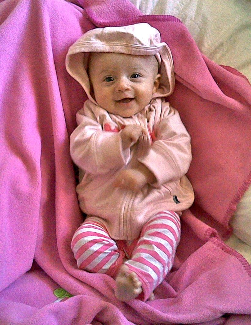 A photo of a small baby lying on a pink blanket with pink and white stripy pants on, and a pink hoodie. She's smiling and is mid-flailing her arms around.