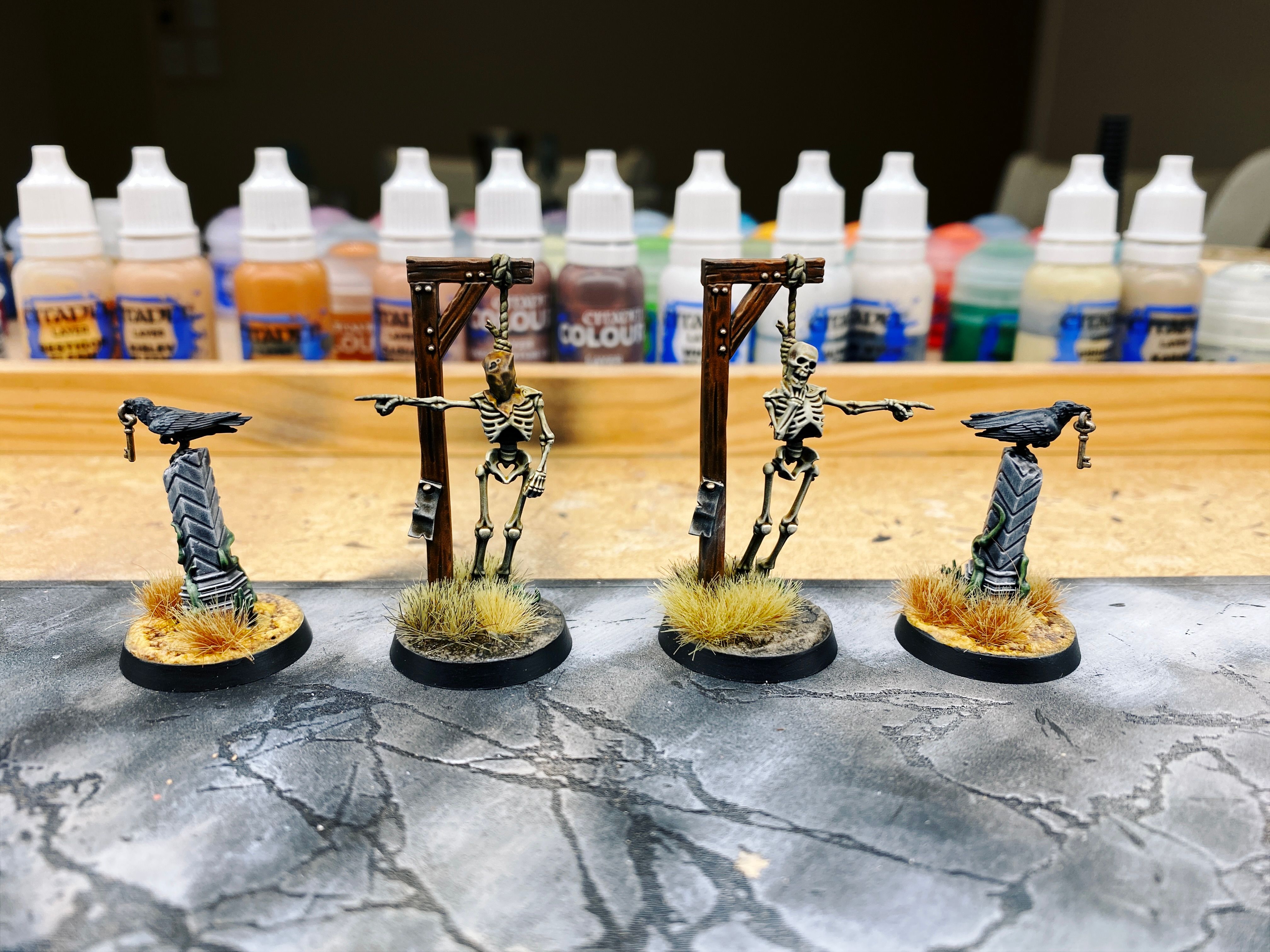 A photo of painted four miniatures. Two are skeletons that have been clearly executed by hanging from fairly hastily-erected gallows but somewhat creepily they both have one arm up and are pointing at something. The bases look like dirt and have some greeny/brown grass tufts on them. The other two are crows that are standing on top of a ruined column, with a key hanging from their beaks. The bases of these look like some sort of yellow stone and have very dry-looking grass tufts on them.