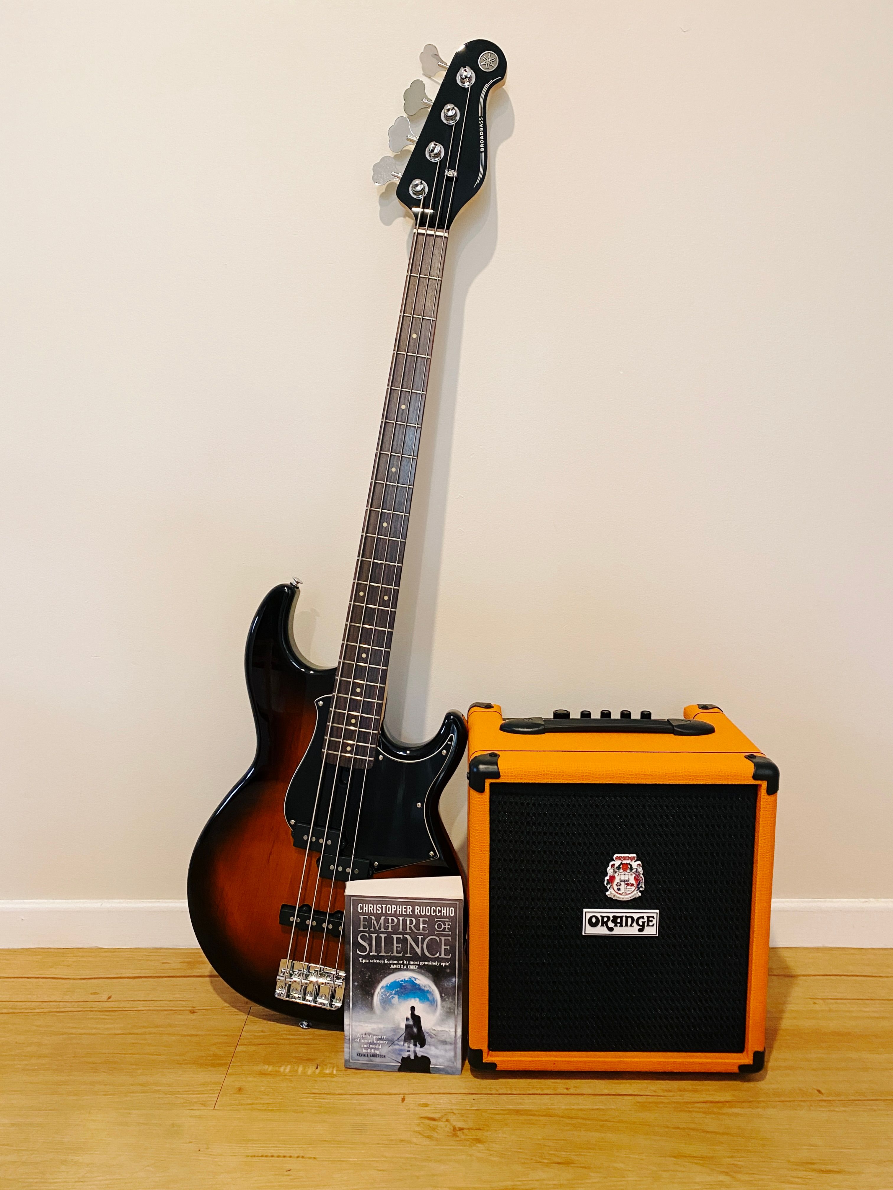 A photo of a small bass guitar amp with a bass guitar leaning against it, and a novel sitting in front. The amp is an Orange Crush Bass 25 and has a black mesh on the front with the rest of it being the customary bright orange colour. The guitar is a Yamaha BB434 and the body fades from black around the edges into a dark rich orange/red wood in the middle. The book is a sci-fi novel "Empire of Silence" by Christopher Ruocchio.