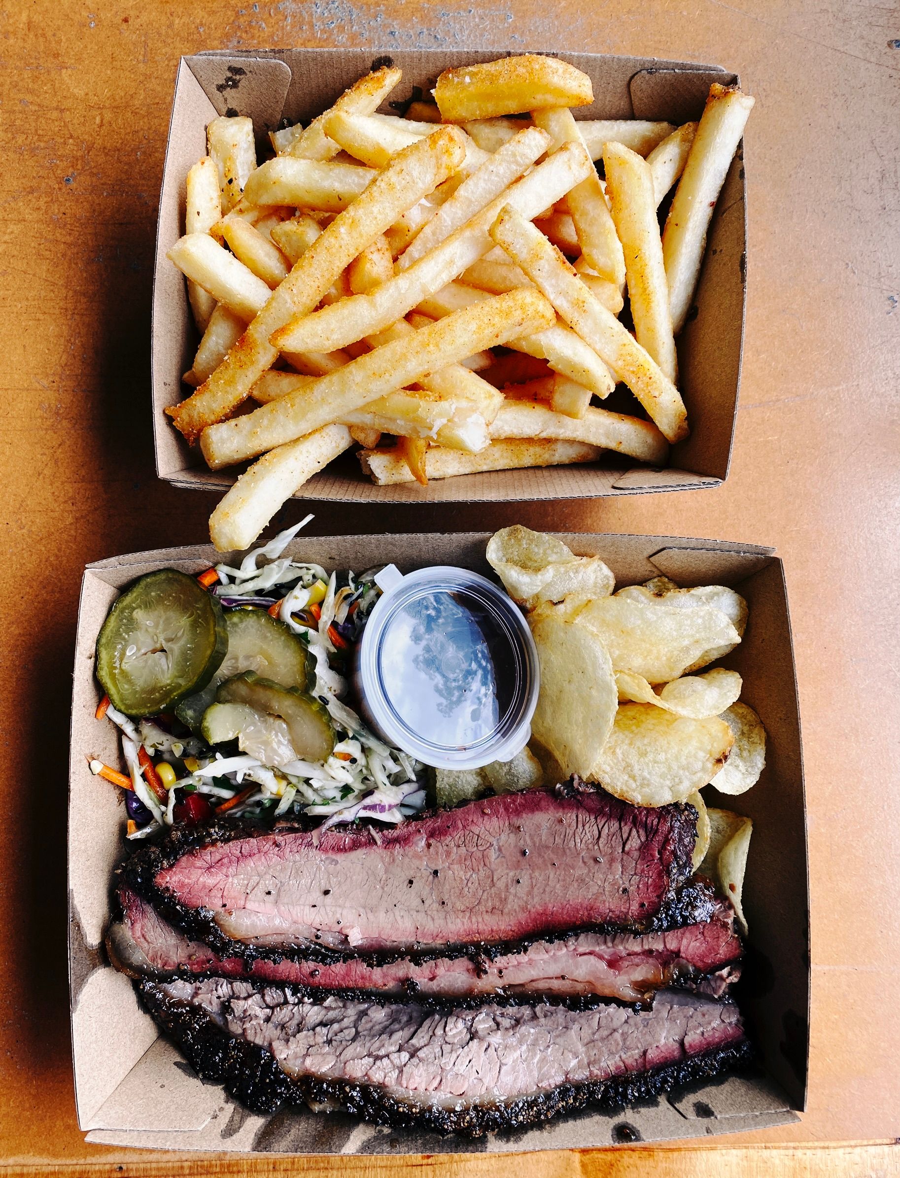 A photo of two cardboard containers, one has three slices of beef brisket along with potato chips, some pickles, slaw, and a container of barbecue sauce, and the other has hot chips in it.