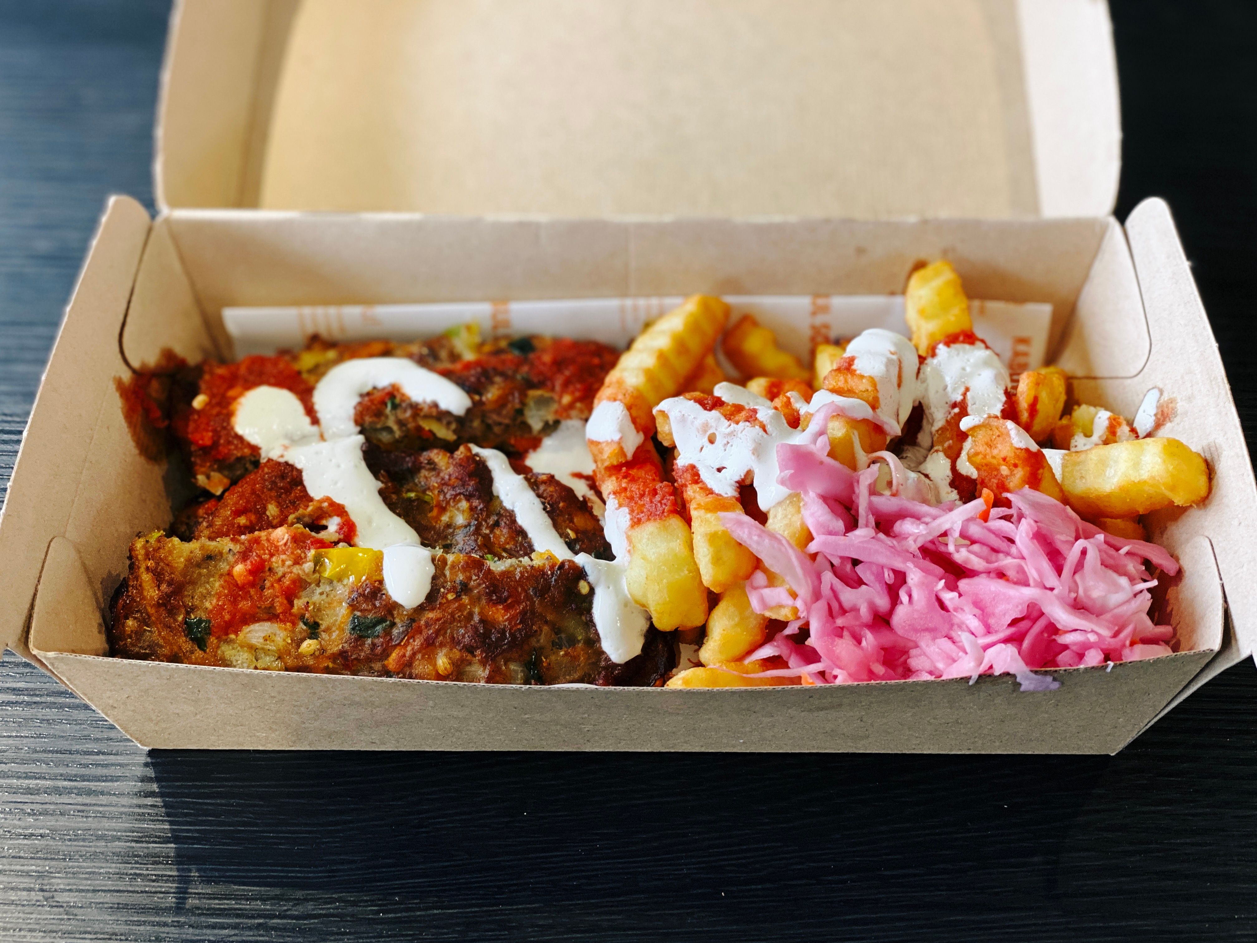 A photo of a takeaway cardboard food container with three pieces of lamb mince with herbs and onions in them, alongside fries drizzled with red chilli sauce and yoghurt, and there's very pink cabbage sitting next to the fries. It looks fantastic and tasted even better.