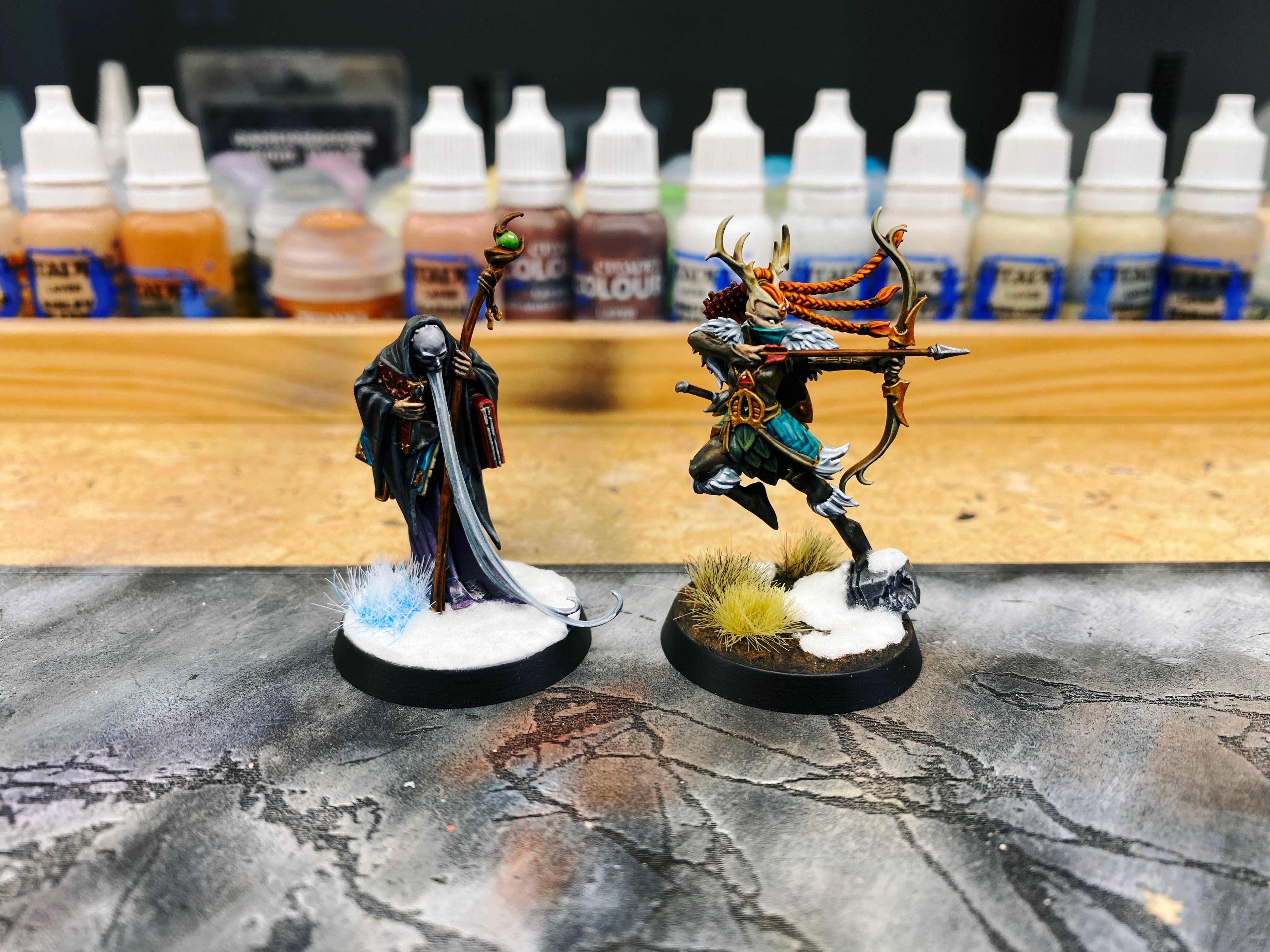A photo of two painted miniatures. One is a wizened old man with an EXTRAORDINARILY long and thin beard that reaches down to the ground, wearing a black cloak and carrying several large books along with a tall wooden staff. Standing next to him is a lithe wood elf all dressed in greens and browns with a pair of antlers on her head. She's in the middle of aiming a bow an arrow and is simultaneously launching herself off a broken piece of stone on the ground. There's snow at the base of the stone and next to it, but the left half of the base is rich earth with grass tufts on it.