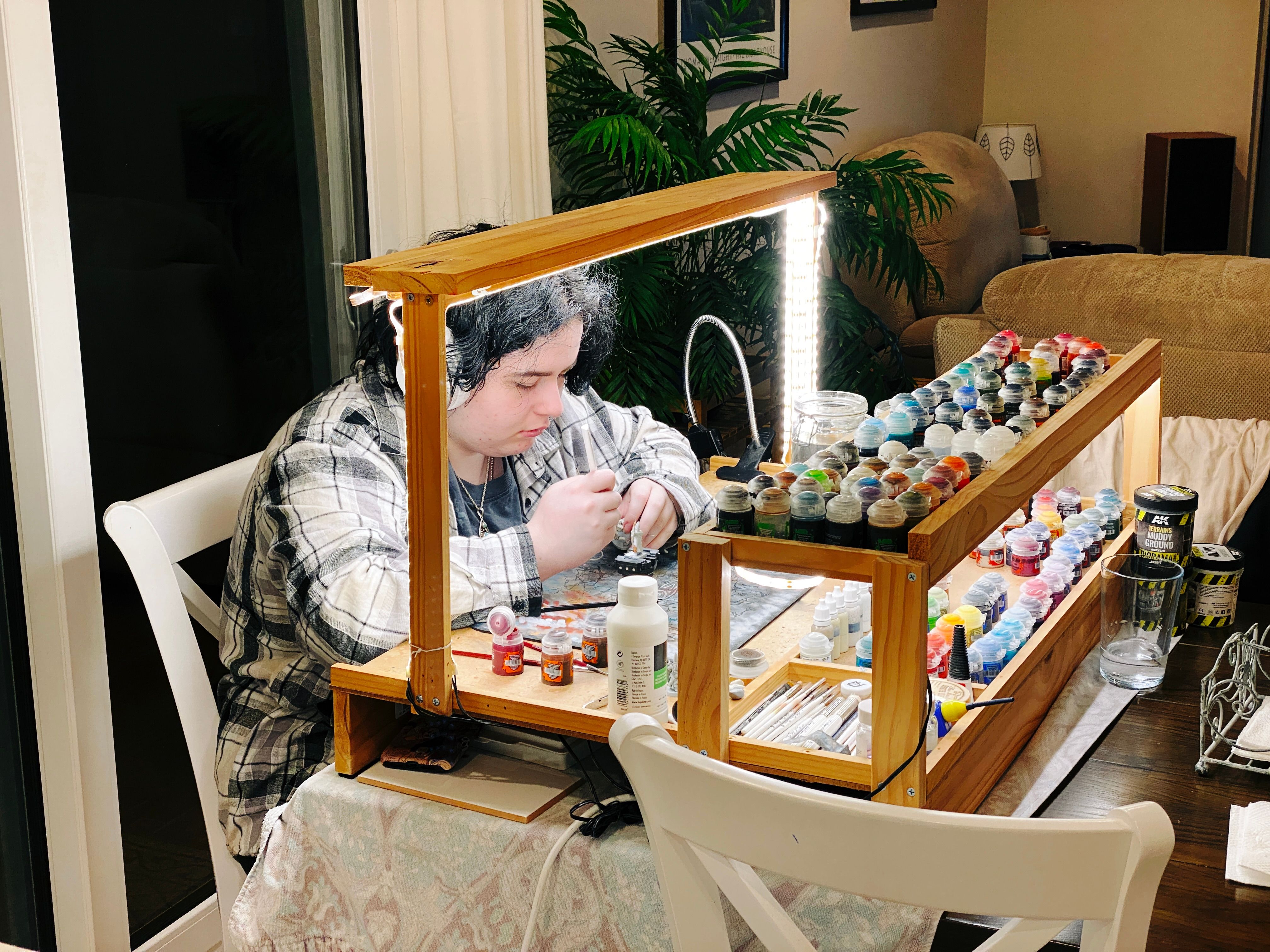 A photo of a white non-binary person with black hair and white headphones on, holding a paintbrush and intently looking at the miniature they're painting. They're sitting at my painting table which has a wooden arch with LED strip lighting in the middle, and two levels of storage for my many many paint pots.