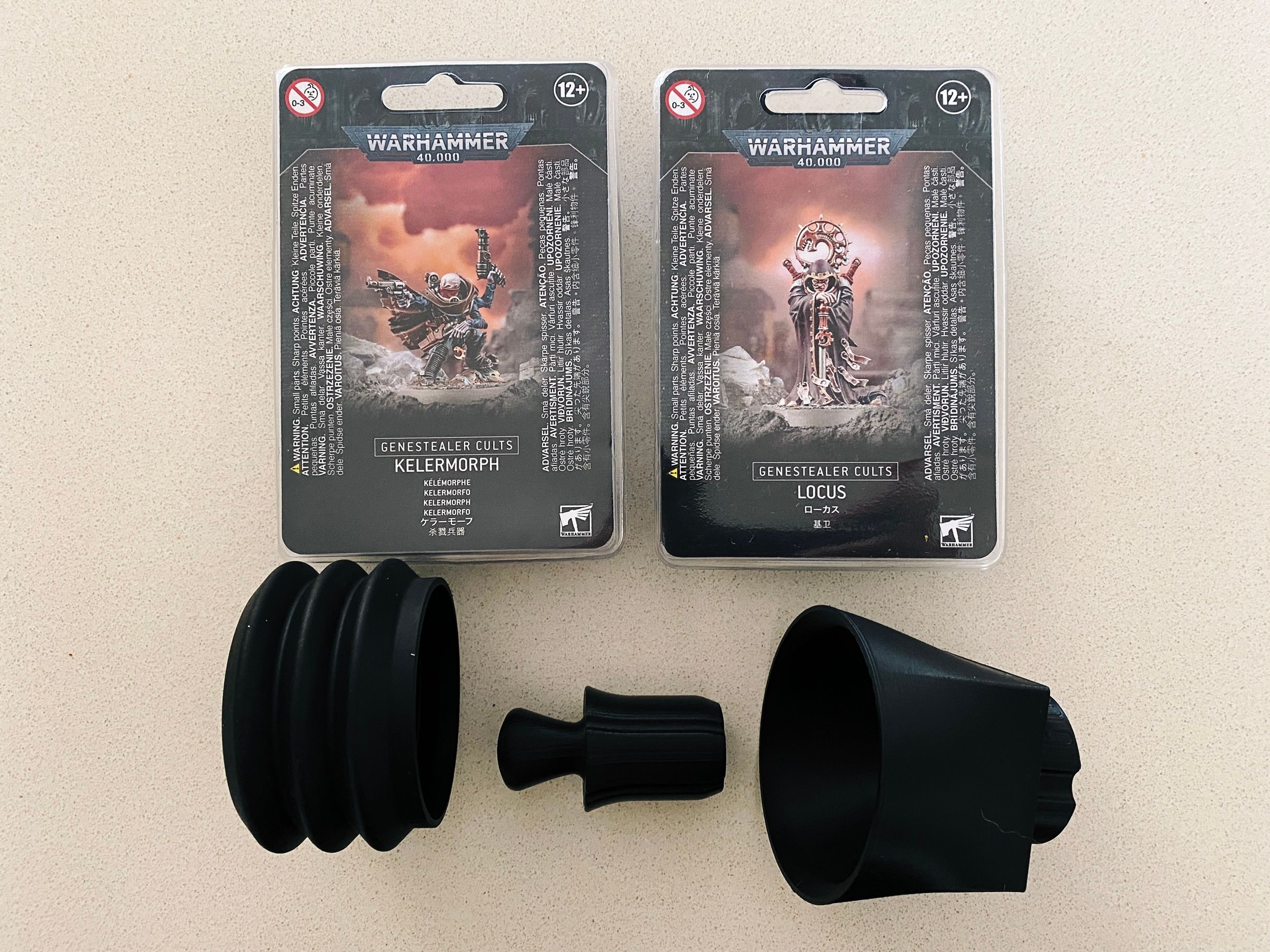 A photo of the boxes of two Warhammer 40,000 miniatures, both are freaky-looking human/alien hybrids (one looks like a gunslinger and the other is standing there looking menacing). Underneath those are three plastic things: a little bellow, a funnel-looking thing, and a small weight.