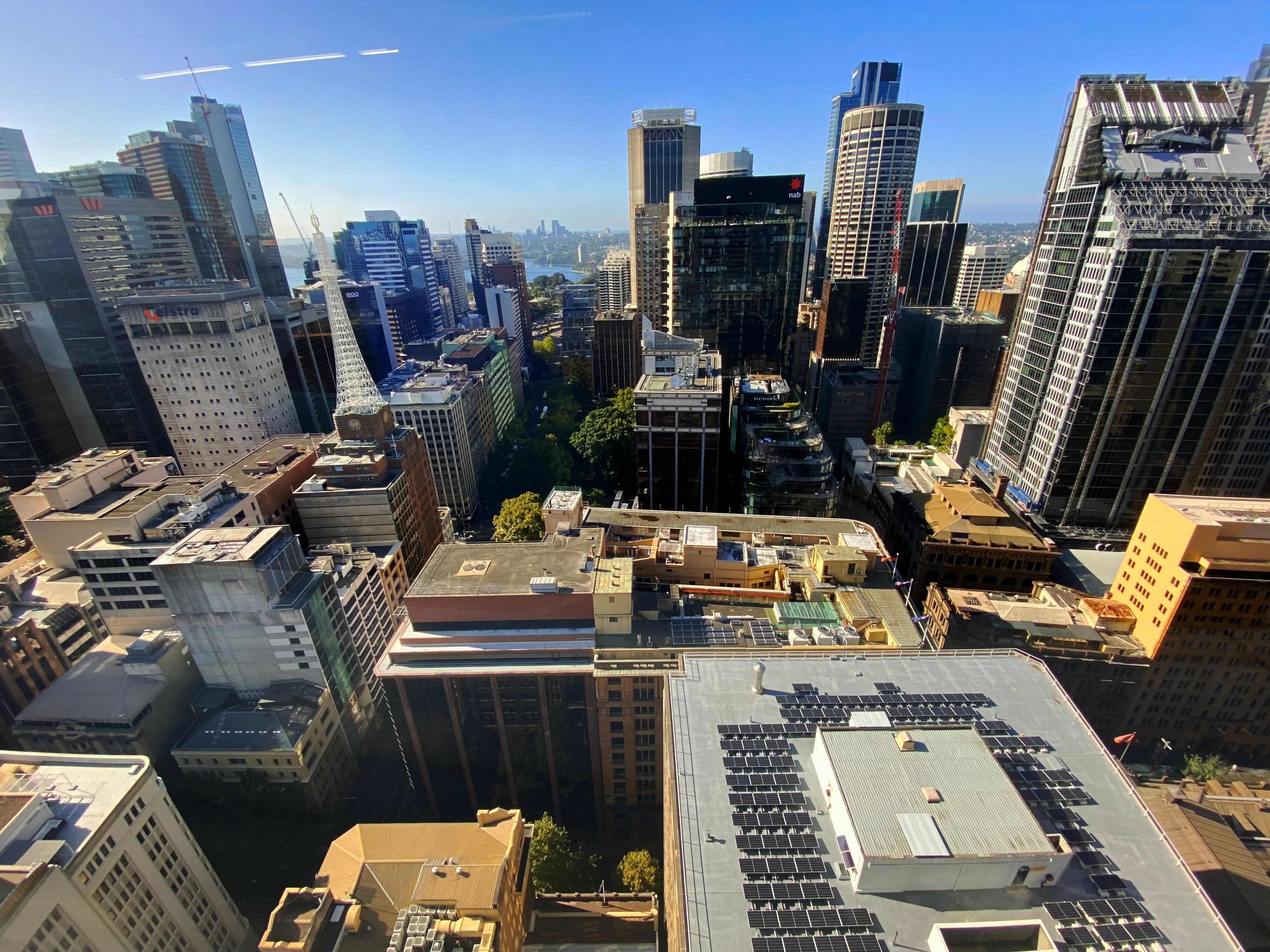 A photo taken from the 28th floor of our office building looking out over Sydney's CBD. Lots of tall buildings, with shorter ones directly below. The sky is a lovely blue colour and what I think is North Sydney is distantly visible through a gap between two of the buildings.