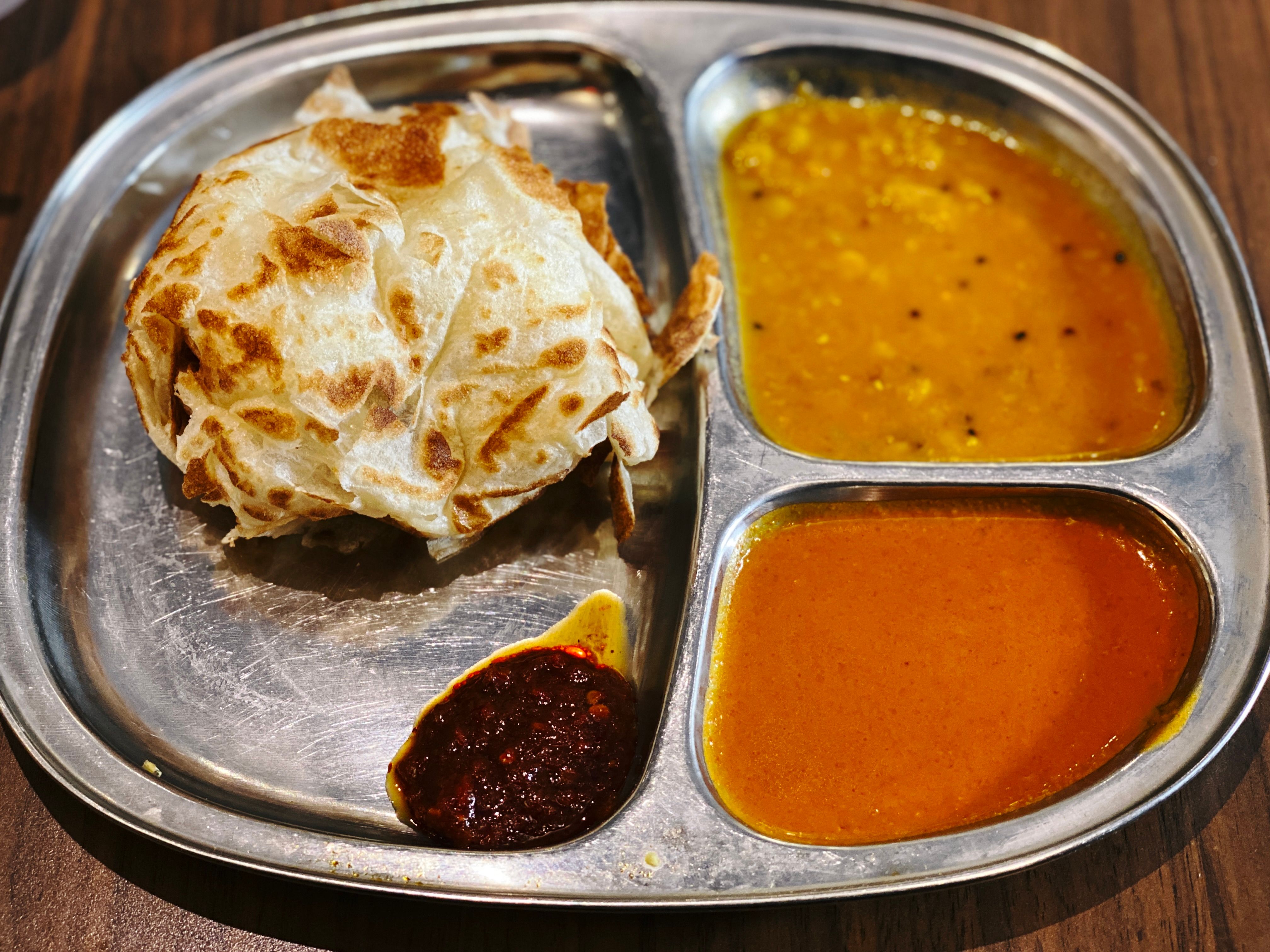 A photo of a three-section metal tray, the left has a roti canai which is a sort of fluffy balled-up pastry thing and a dollop of sambal sauce, and the other two sections have two different types of curry sauce in them.