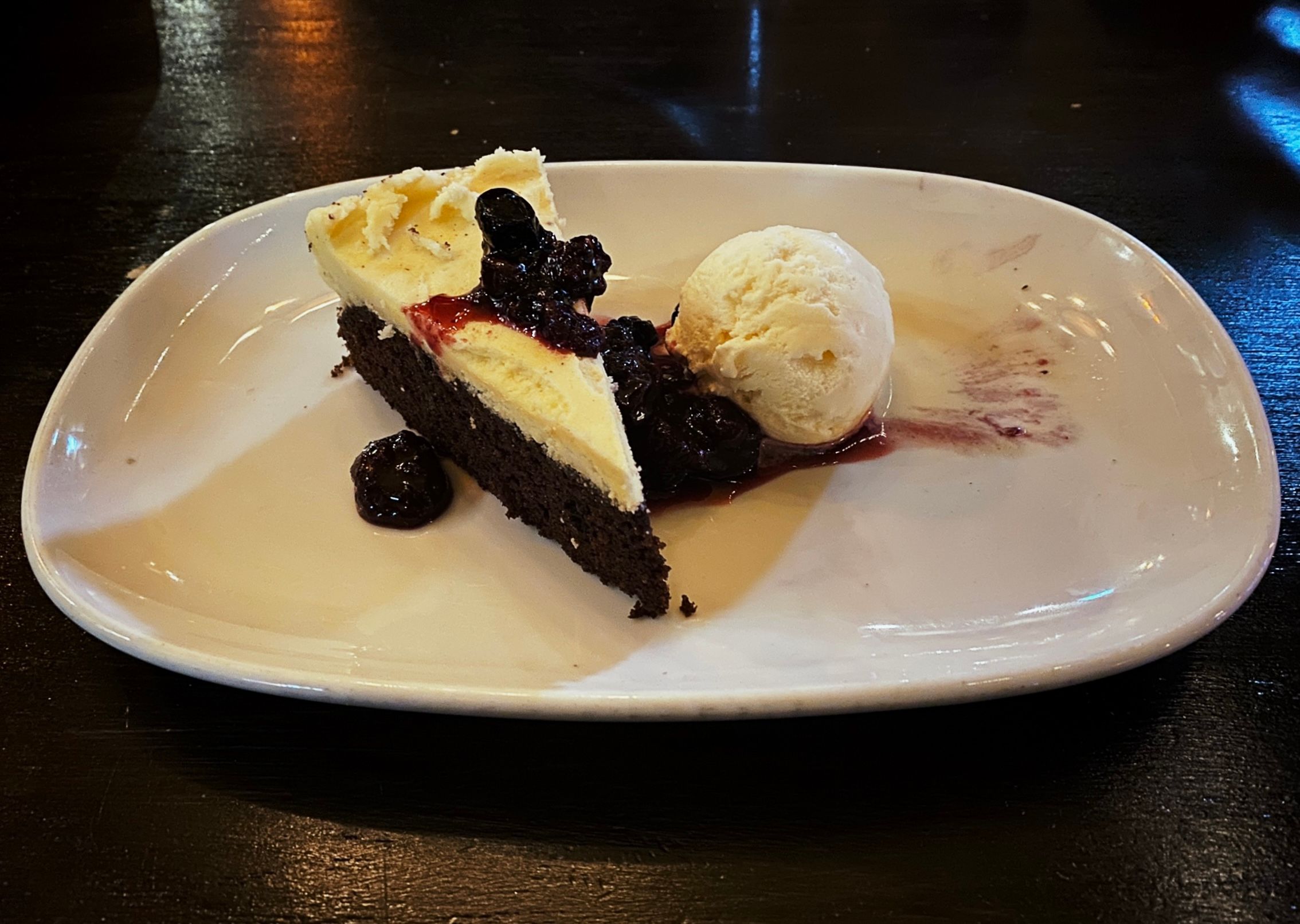 A photo of a slice of dark chocolate cake with white icing on top alongside some berries and a scoop of vanilla ice cream.