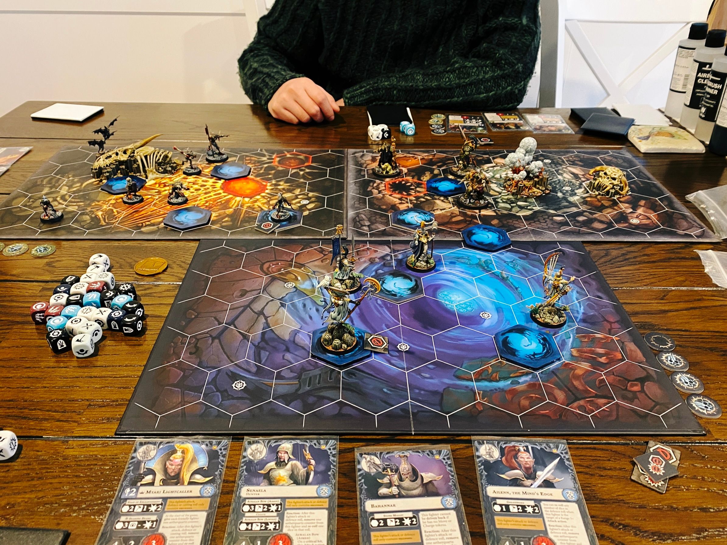 A photo of a Warhammer Underworlds game board setup. There's three individual boards, two are have the artwork underneath looking like deep within a cave and the third looking under it's an underwater cave with lots of blues. At the front closest to the camera are four lithe and elegant aelves, the far side has three diseased Nurgle warriors, and the left has seven gore-covered  ghouls.