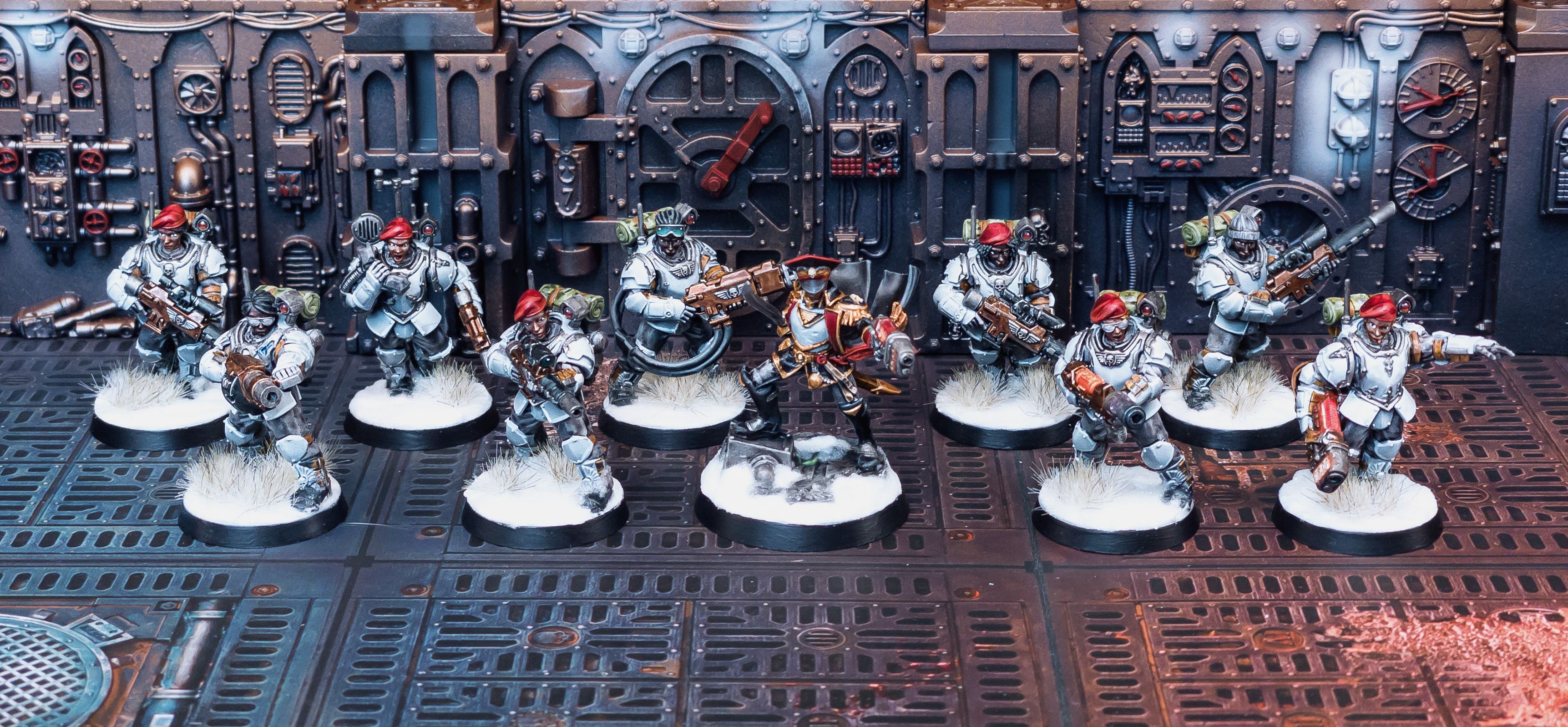 A photo of ten Astra Militarum Tempestus Scions (basically regular humans with guns in armour). The leader is in the middle and standing on a piece of industrial machinery aiming a plasma pistol and has her black cape billowing out dramatically behind her. They others are all wearing big blue-grey armour and are wielding various guns, they've all got black or brown skin, and they all look extra bad-ass because they're wearing red berets and a couple of them have sunglasses on. The bases are done so they all look like they're standing in snow.