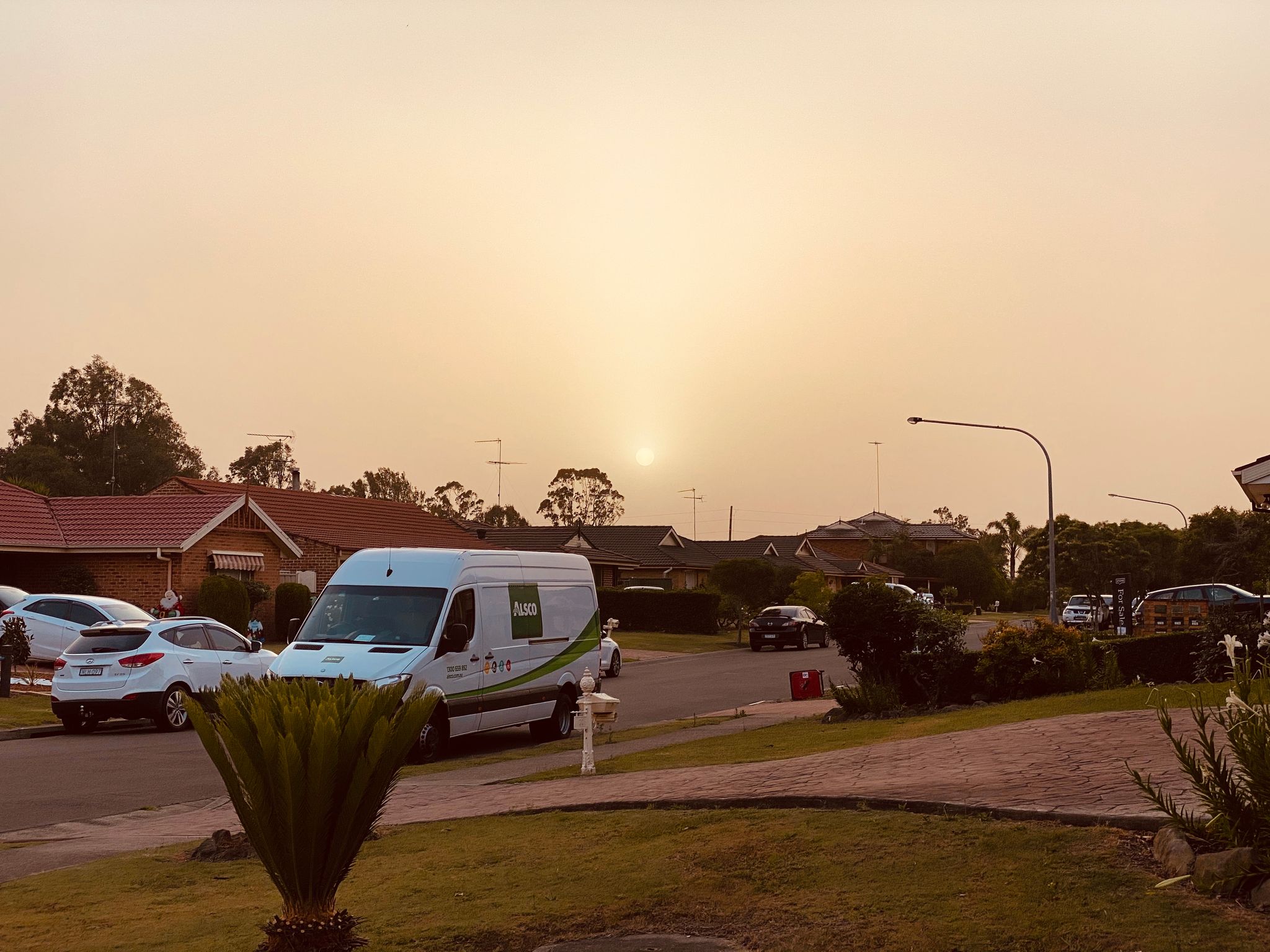A view down a suburban street at sunset, the sky is brown-tinged and the sun is a disc that's able to be looked at with the naked eye because there's so much dust in front of it.