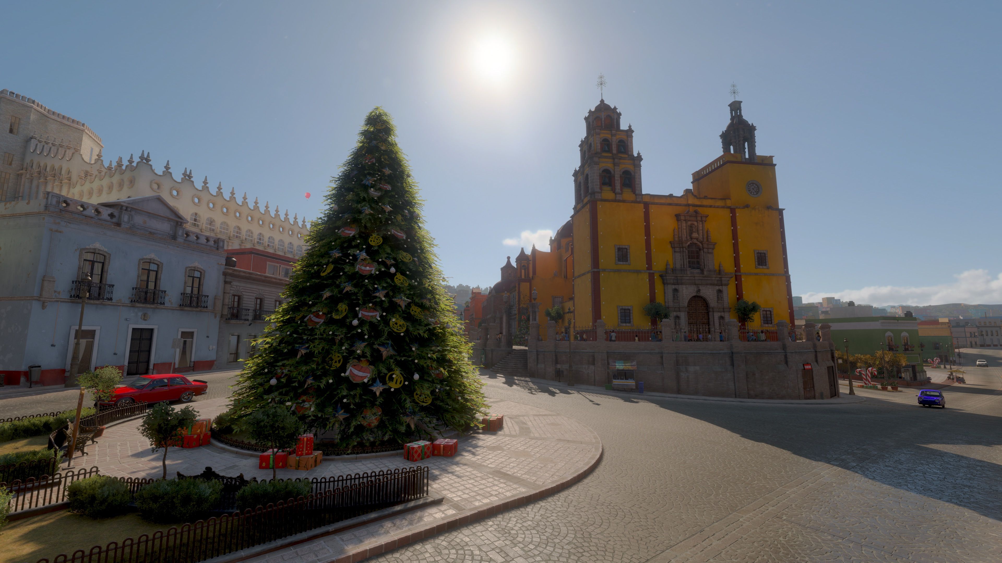 A screenshot from Forza Horizon 5, it's a sunny day with cobblestone streets and obviously-Mexican buildings, and there's a massive Christmas tree in the middle the section where a road is splitting apart. On the other side of the road is a huge yellow building that I think is a church.
