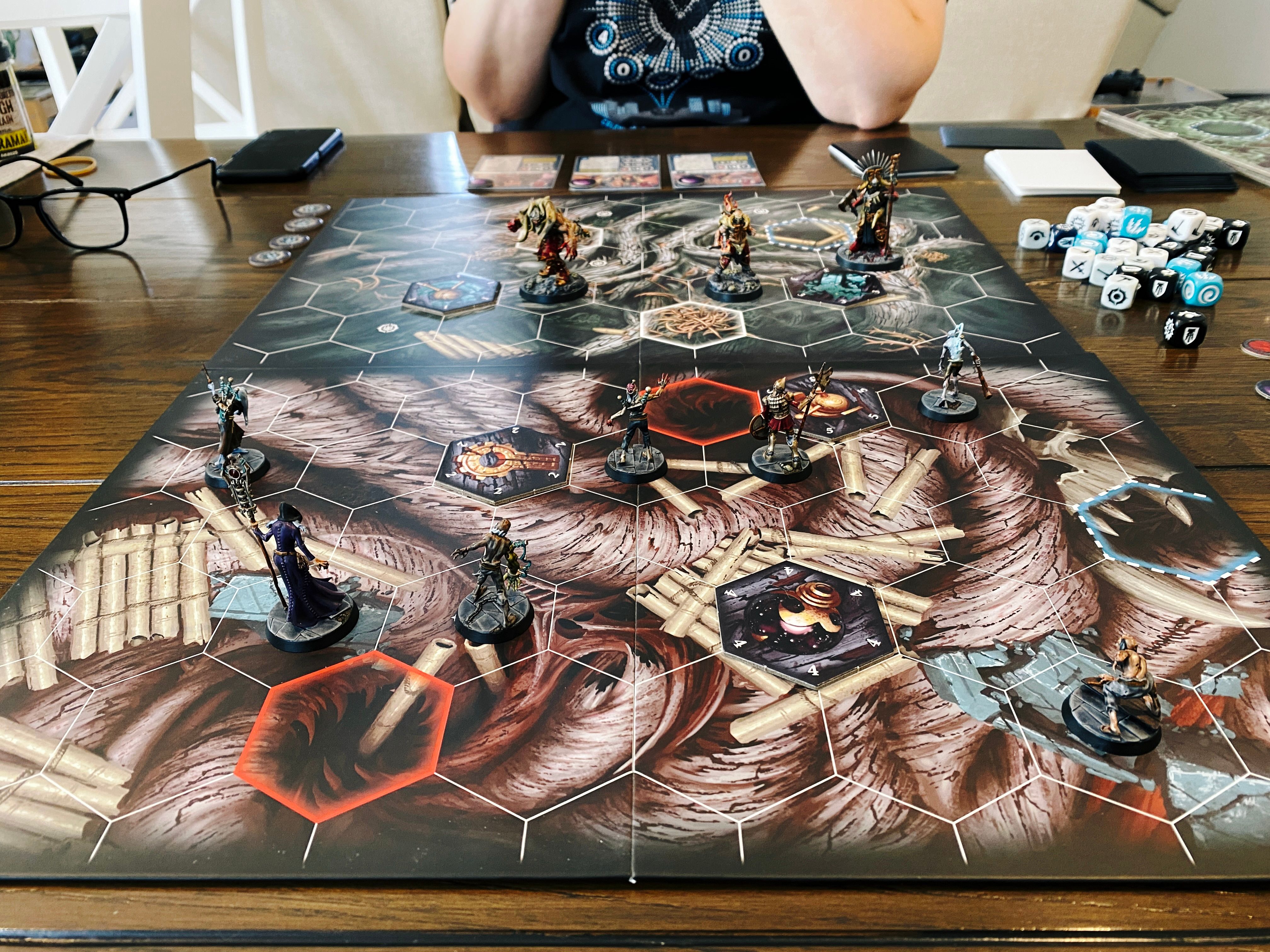 A photo of a Warhammer Underworlds game board. It has hex tiles on it, and the artwork looks like the bottom of a forest with gnarled roots everywhere. Closest to the camera are the Exiled Dead, a warband of seven miniatures consisting of six zombies and the zombiemaster Deintalos. Four of the have very Frankenstein-esque metal poles coming out of them like they're being animated by electricity, the fifth is more upright and has armour and a halberd, and the sixth is crouched down low to the ground. Deintalos is wearing a flowing purple robe and carrying a staff. On the other side of the board are the Gorechosen of Dromm, a three-fighter warband consisting of very large meaty-looking warriors in large red armour.