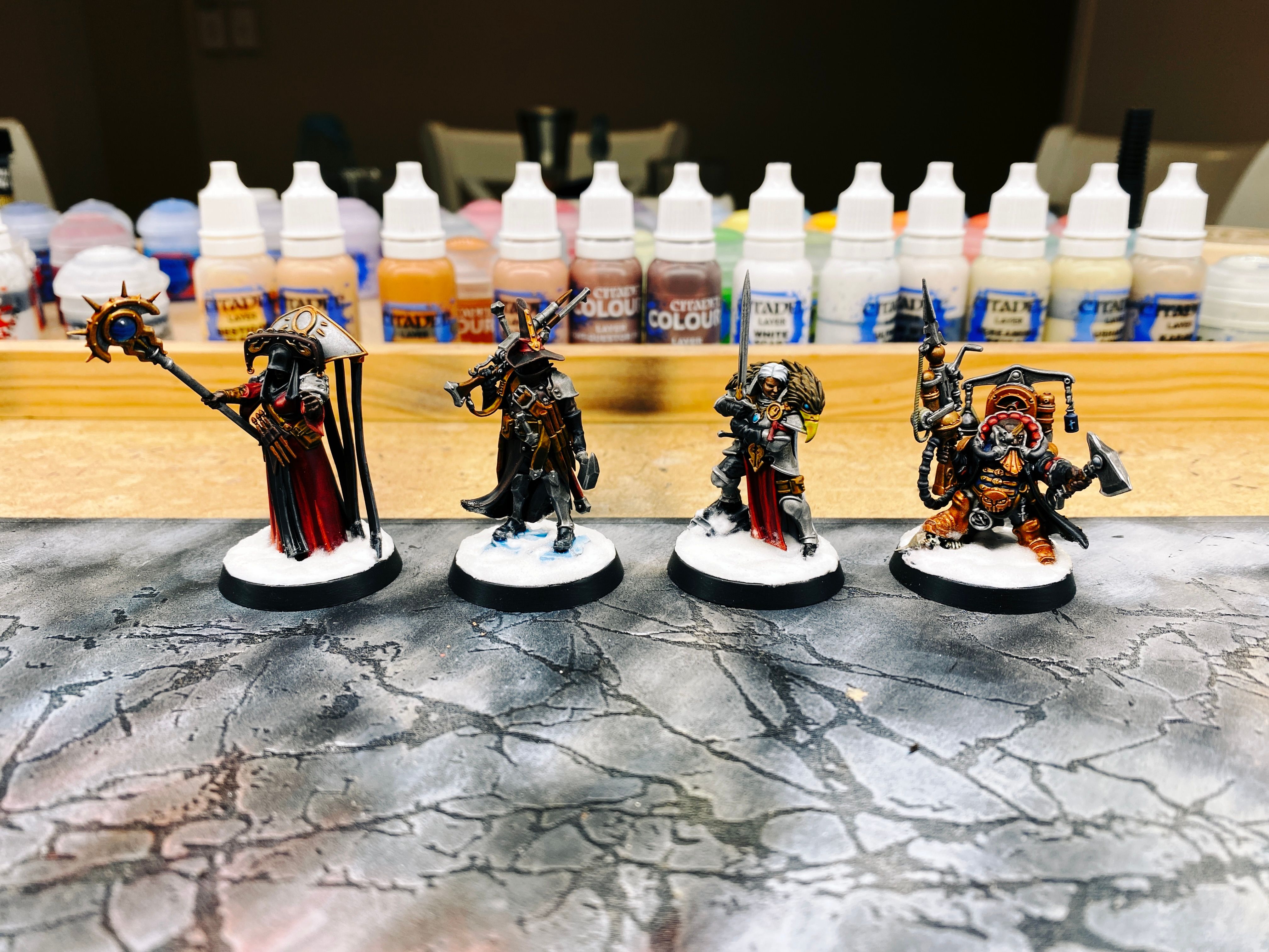 A photo of four painted miniatures:
- A vampire hunter with the standard pointy hat and cool dark brown leather cloak holding a shotgun-looking thing, and with many wooden stakes about his person.
- A heavily-armoured warrior woman with white hair holding a large sword in front of her, with the head of a gryphon draped over her shoulders.
- A dwarf with steampunk-looking armour holding an axe and a steampunk-looking gun.
- A priest in long deep rich red robes with a black hood on and a big elaborate, uh... metallic thing on her back that a bunch of strips of fabric are hanging from. She holding a staff and has her hand out like she's about to cast a spell.