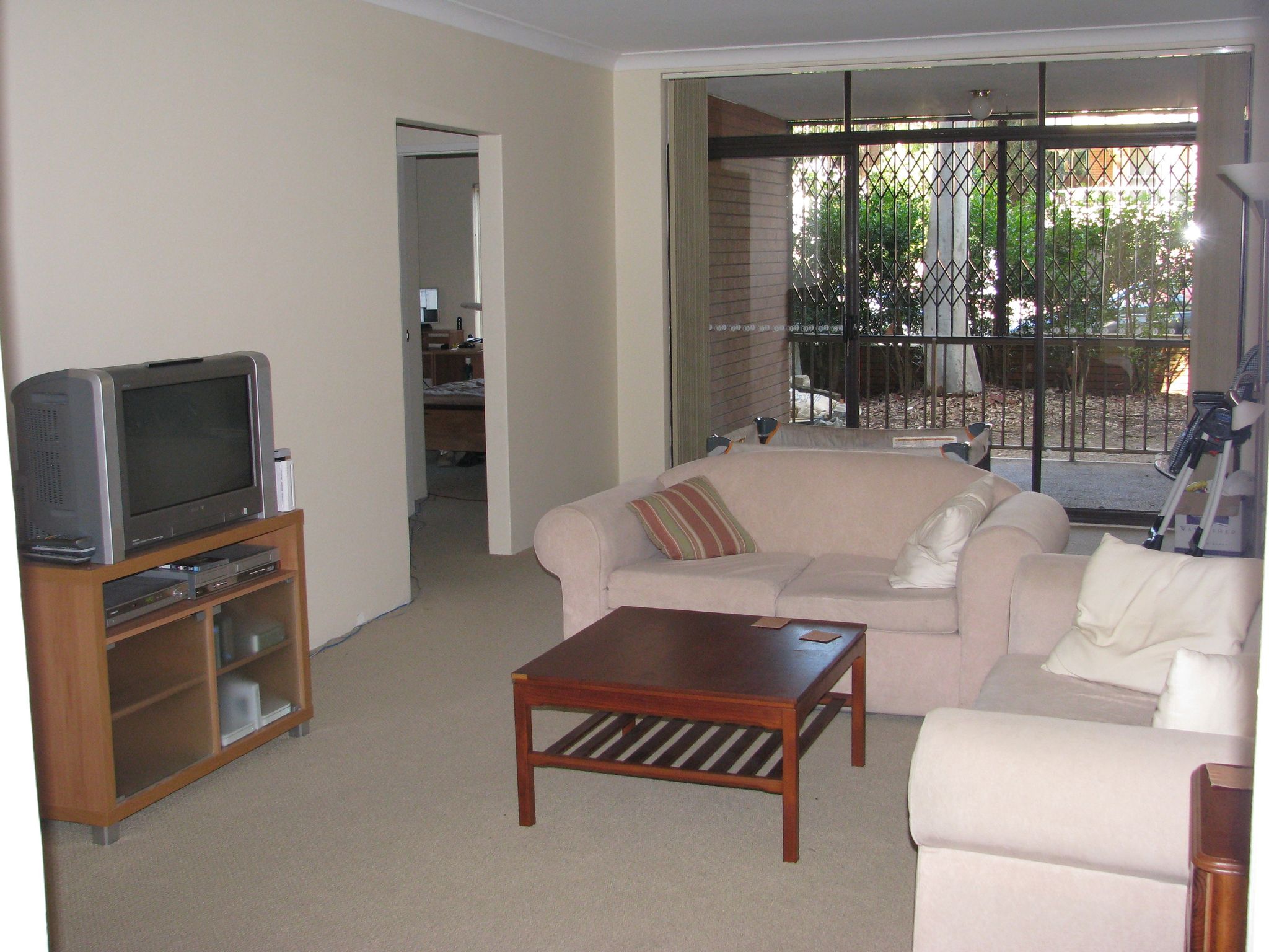 A photo of the loungeroom inside a ground-floor unit. There's a small balcony facing where the camera is pointing, with horrible security bars over the gap between the railing and the ceiling. Inside on the left wall is a CRT TV on top of a wooden TV stand with stuff inside it. Facing away from the balcony is a two-seater lounge, and to the right of that facing the TV is another two-seater lounge of the same sort. There's a low square brown wooden coffee in between the lounges.