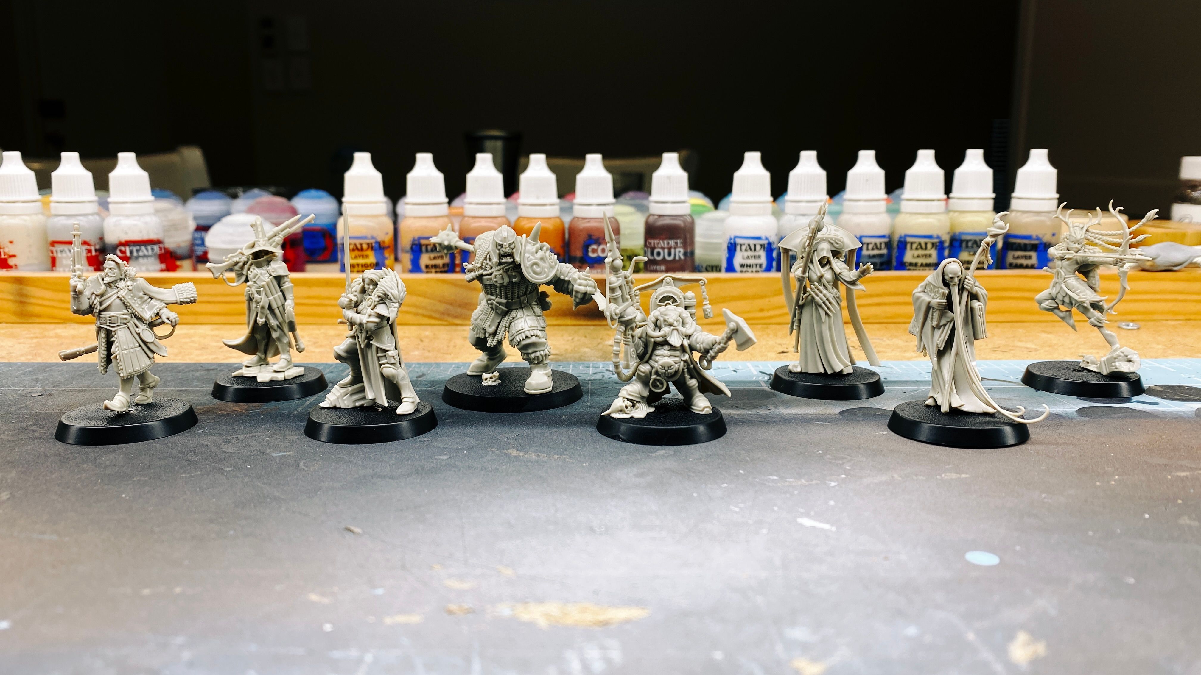 A photo of eight assembled but unpainted miniatures in bone white. We have:
- An aristocratic-looking fellow with a goatee and holding a old timey-pistol and a sword, mid-swagger.
- A vampire hunter with the standard pointy hat and cool cloak holding a shotgun-looking thing, and with many wooden stakes about his person.
- A heavily-armoured warrior woman holding a large sword in front of her, with the head of a gryphon draped over her shoulders.
- A HUGE ogre that's a good couple of heads taller than anyone else, with a massive mace that he's about to swing.
- A dwarf with steampunk-looking armour holding an axe and a steampunk-looking gun.
- A priest in long robes with a hood on and a big elaborate, uh... thing on her back that a bunch of strips of fabric are hanging from. She holding a staff and has her hand out like she's about to cast a spell.
- A wizened wizard who's hunched over in a big robe and has a like skull mask on and a MASSIVE long thing beard, he's holding a staff and clutching a very large tome under the other arm.
- And lastly, a elf with a bow who's mid-leap and also mid-shot, with her braids out behind her. She's got a bandana over her mouth and nose and her headdress is antlers.