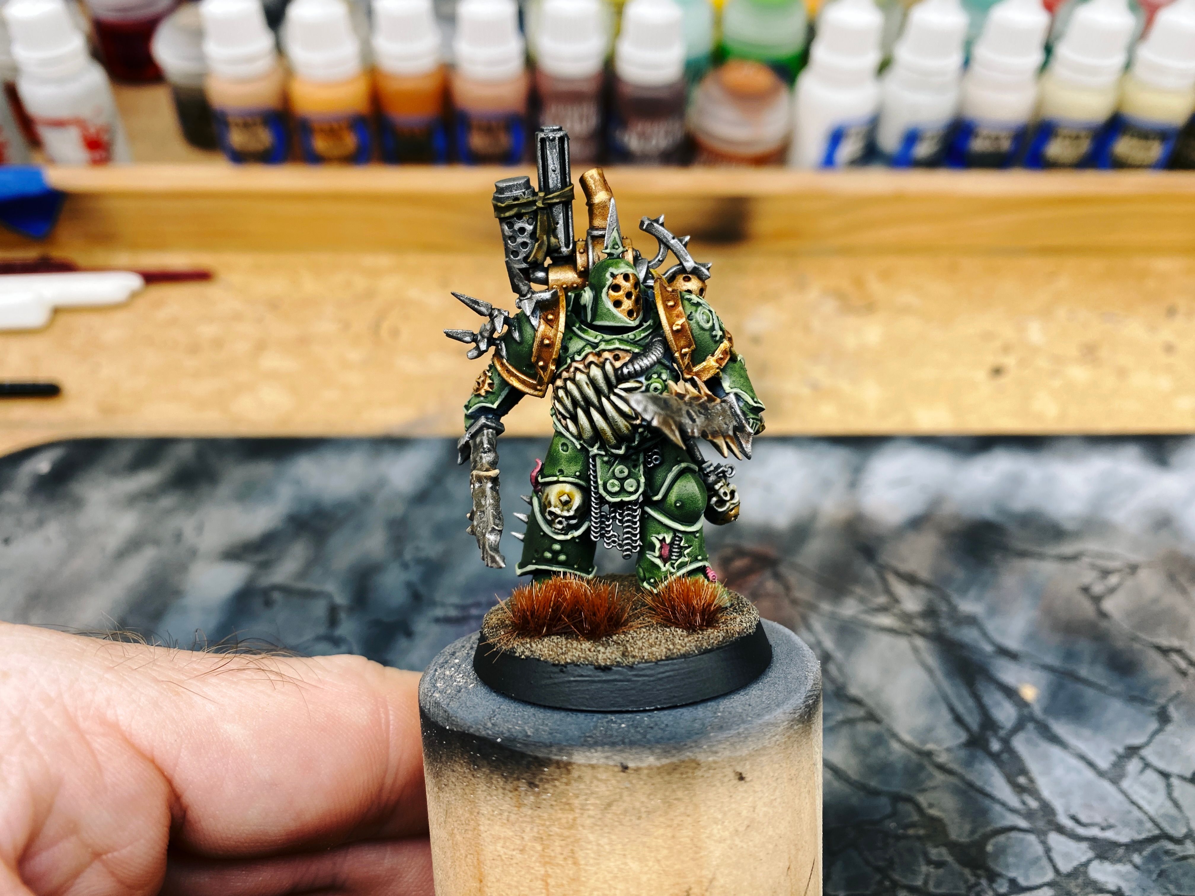 A photo of a Plague Marine from Warhammer 40,000. He's in huge khaki green standard-issue huge Space Marine armour with bronze-coloured edging in places, but because he's one of Nurgle's chosen he's all corrupted and mutated: there's a GIANT mouth with huge teeth right across the entirety of the chest and abdomen of his armour with a metal pipe coming from his shoulder and going into the side of the mouth, and the two very large knives (honestly, they're more like short swords) he's wielding have skin-coloured spines growing out of them in places.