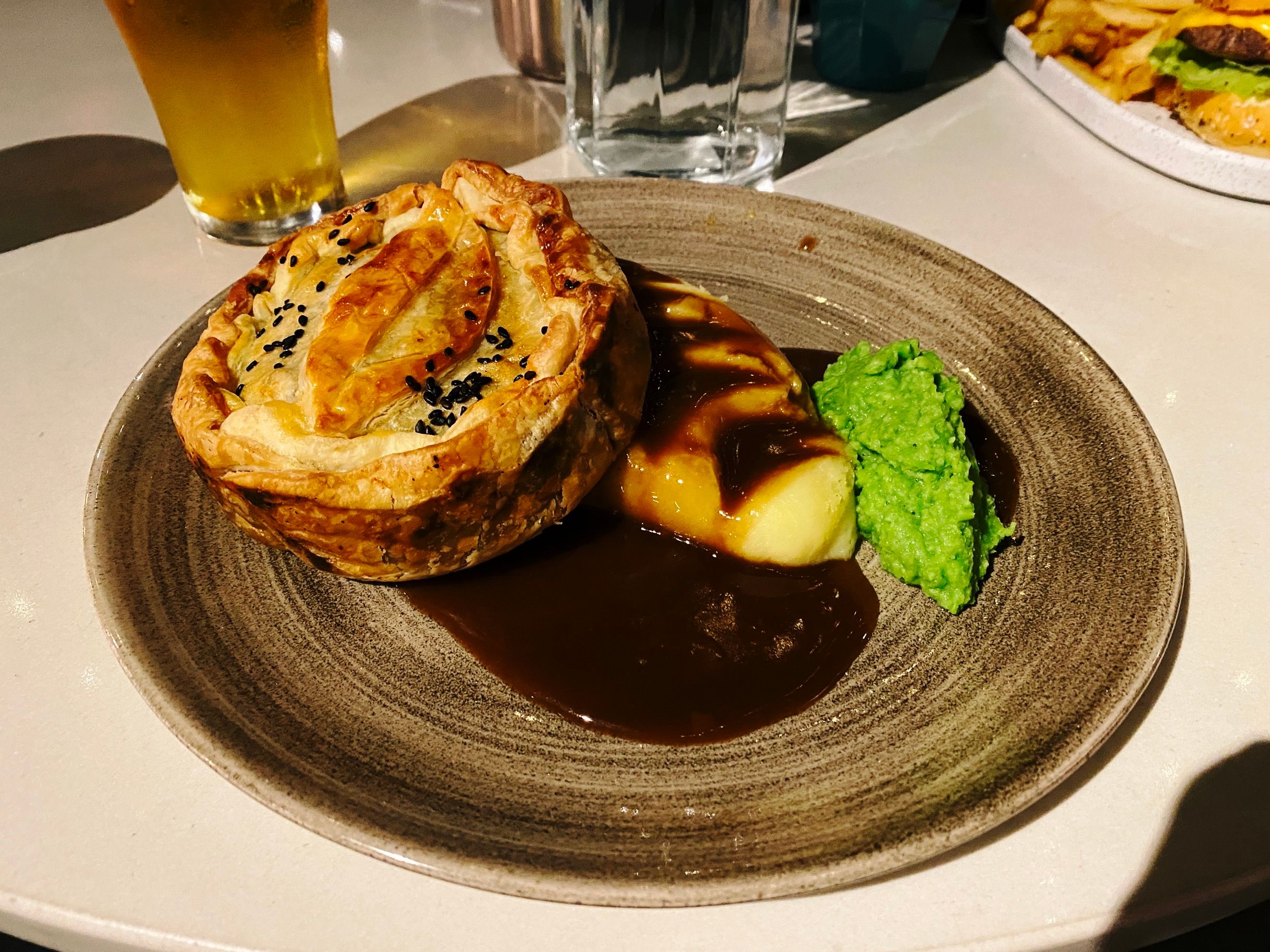 A photo of a pie sitting on a plate along with mash that's covered in gravy and a dollop of mushy peas.