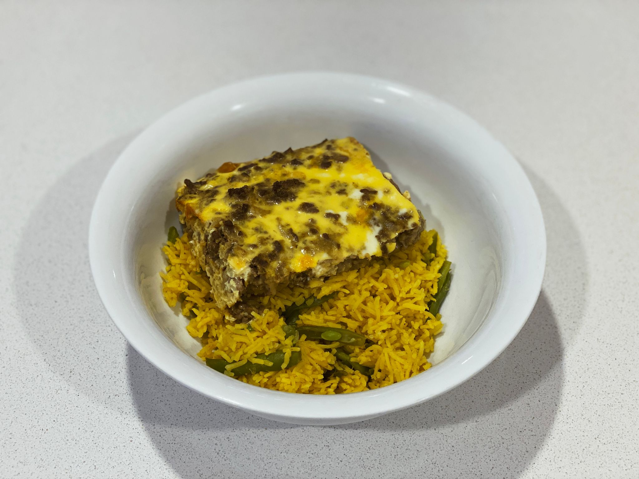 A photo of a quarter of the bobotie sitting in a white bowl on top of yellow rice with green beans in it.
