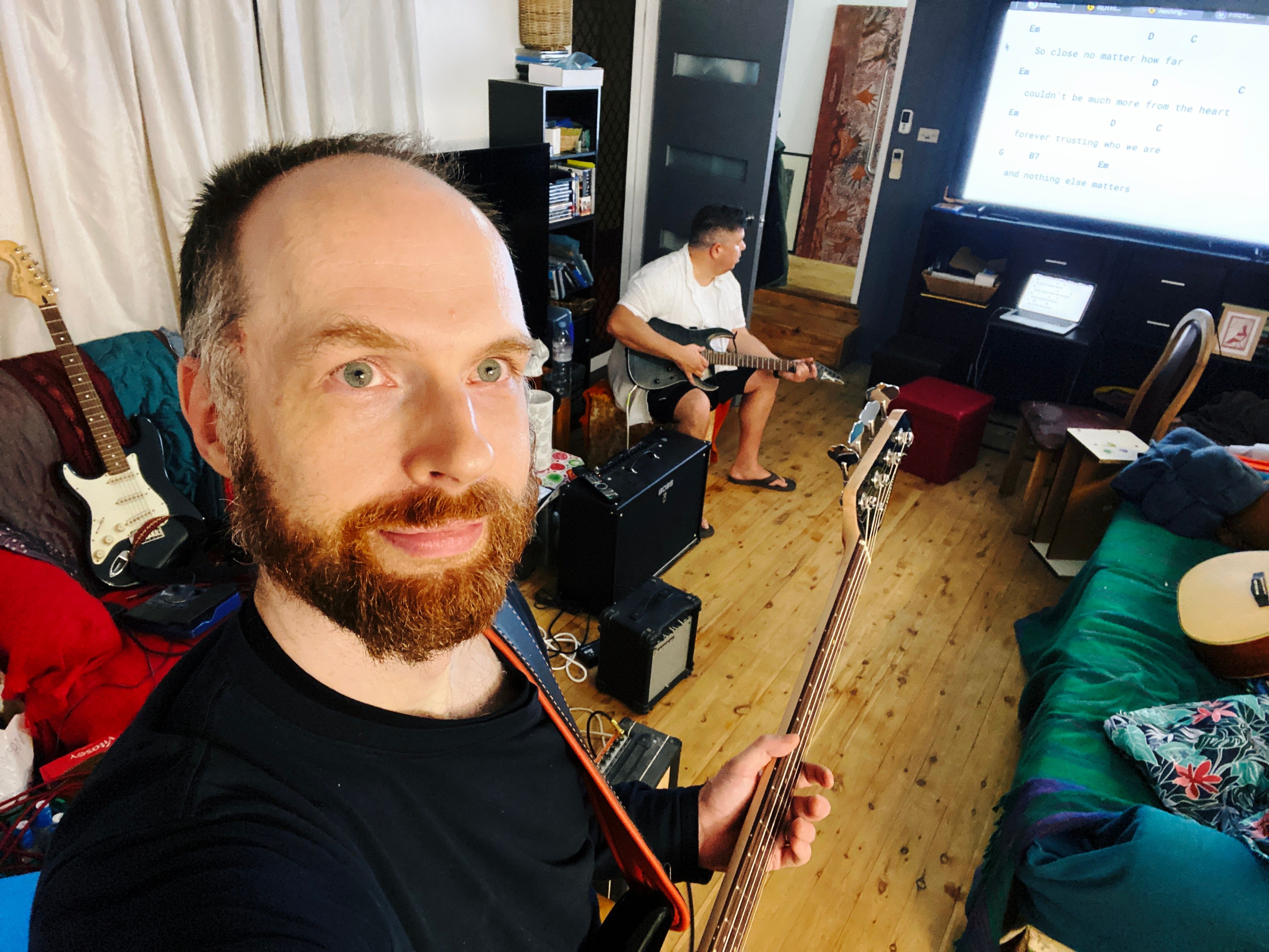 A photo of me, a white man with an increasingly-scruffy-looking red beard, holding a bass guitar and smiling at the camera. My friend is sitting behind me holding a guitar and looking at a large TV where the basic chord progression for Metallica's Nothing Else Matters.