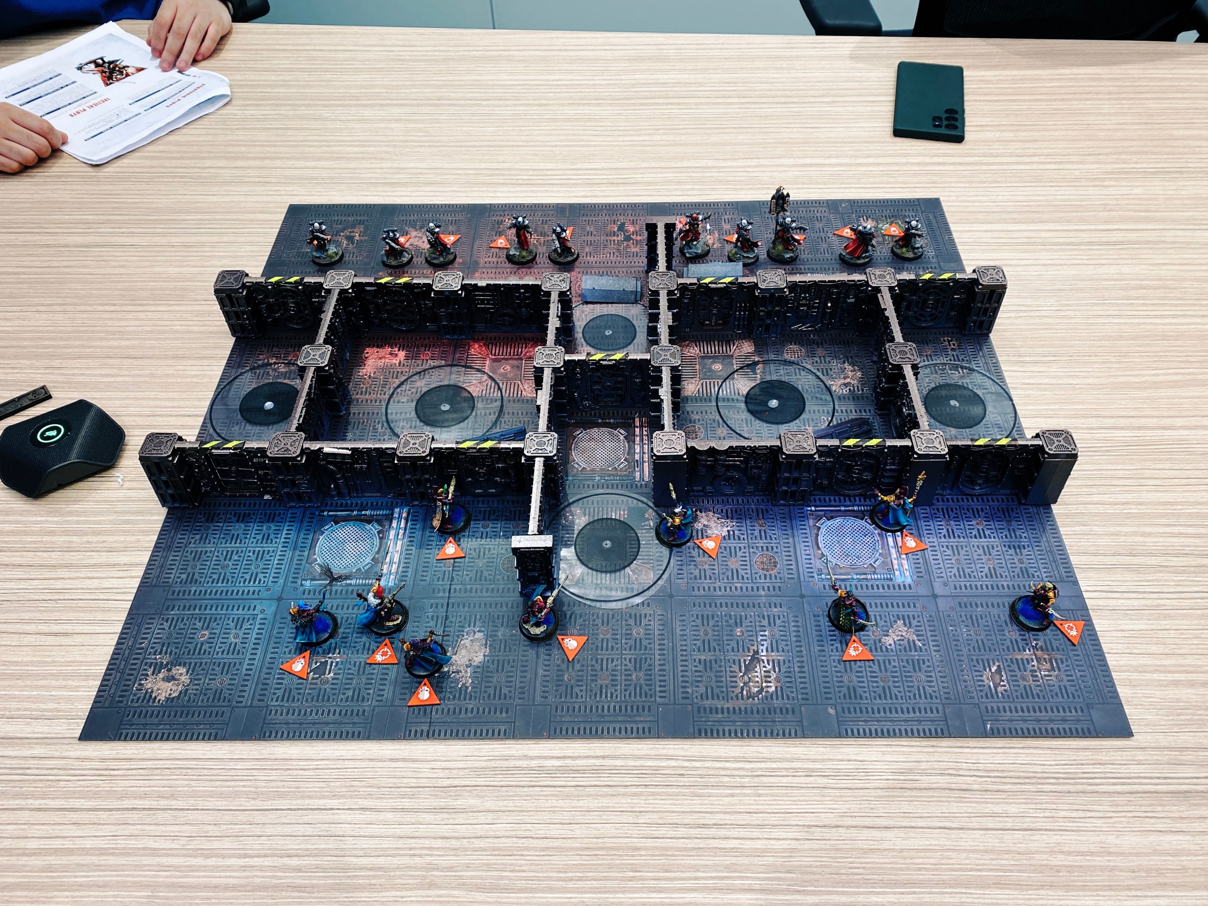 A photo of a game of Warhammer 40,000: Kill Team. The board looks like the metal of the inside of an ancient steampunk starship, and the terrain is a bunch of walls all connected together in the same steampunk style to form number of interconnecting rooms. Closest to the camera are my forces, the Aeldari (think space elves) in elegant fuchsia-coloured armour, and facing them on the other side of the board are ten heavily-armoured human warrior women in black armour and red cloaks, all with striking white hair.