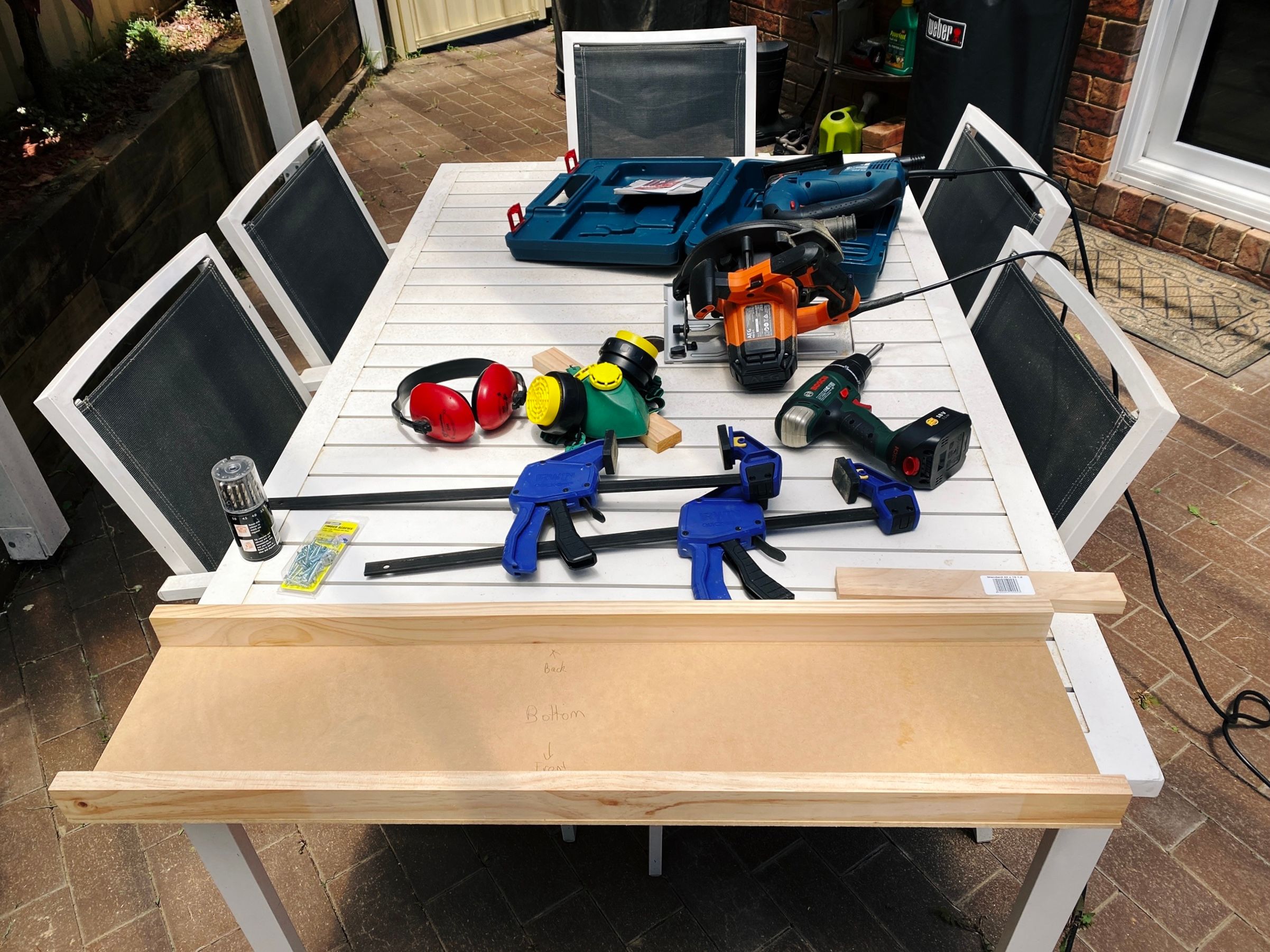 A photo of an white metal outdoor table with several pieces of wood in various states of being cut on it, along with a circular saw and a jigsaw, as well as earmuffs, and a mask,
