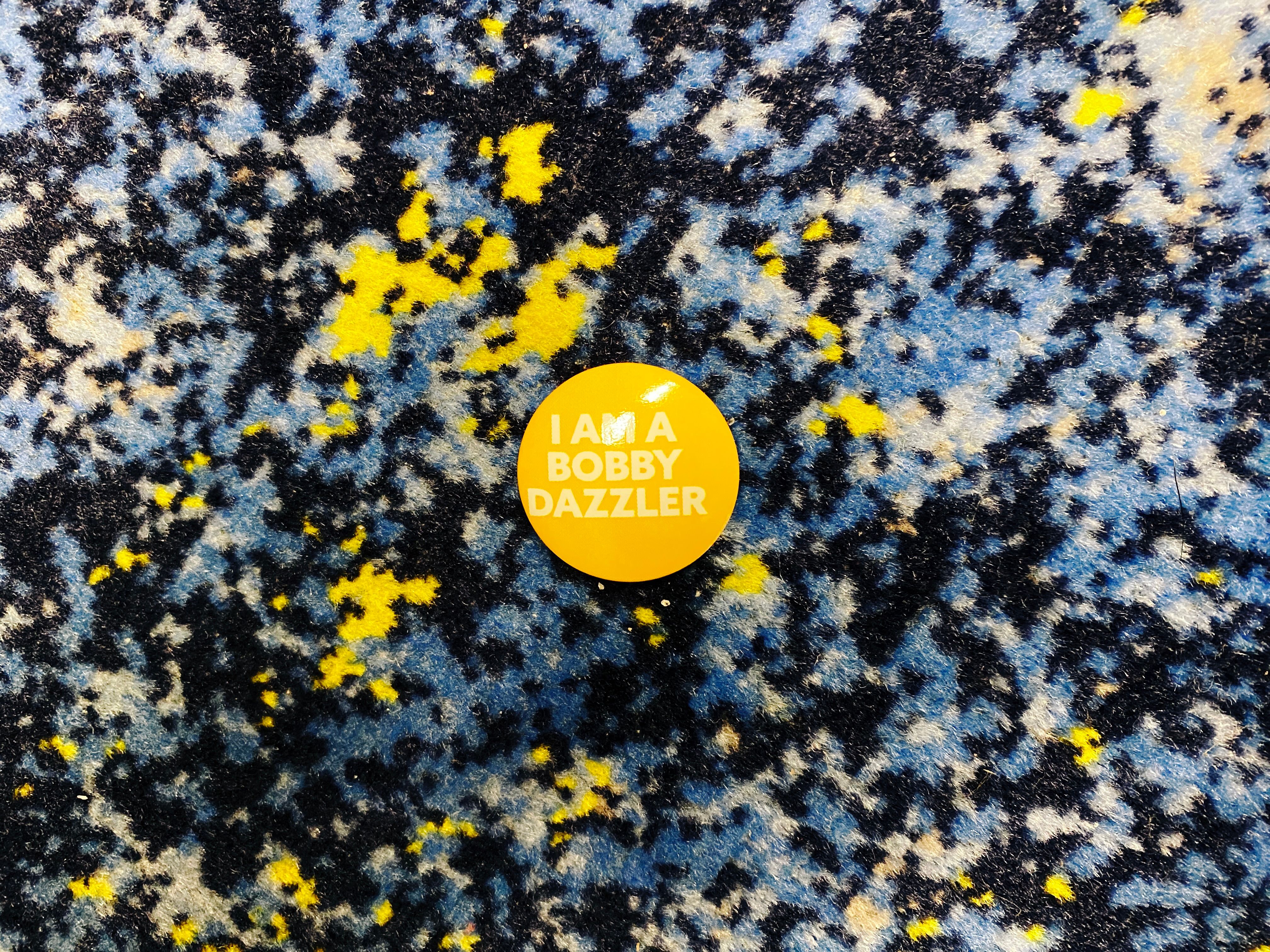 A photo of a small round yellow pin, the sort you'd attach to a lapel or similar that says "I am a Bobby Dazzler" on it.