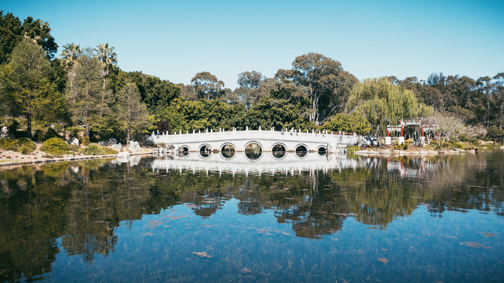 A photo of a white Chinese-style low stone bridge crossing over a lake. There's very little wind so there's a good reflection of the bridge on the surface of the water.