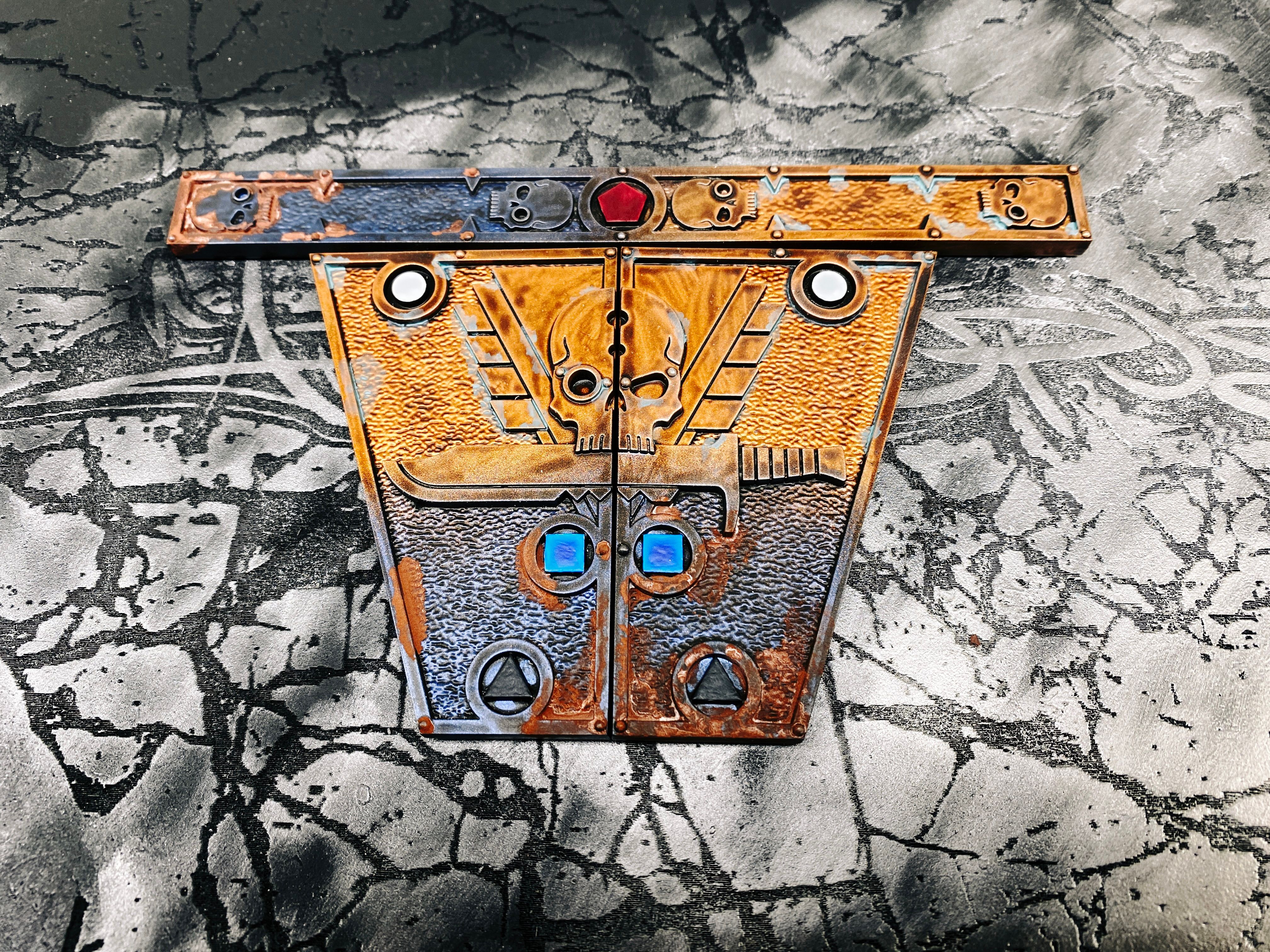 A photo of the three measuring gauges from Warhammer 40,000: Kill Team. One is long and thin with stylised skulls and a red pentagon. The other two are two halves of an isosceles trapezoid with a white circle, blue square, and black triangle on three of their edges, and half of a large skull with a knife under it on either side. All three pieces fade from a dark metallic blue to a bright copper colour, on the dark blue side I've added rust pigment so it looks like it's been rusting, and on the copper side I added a verdigris effect.