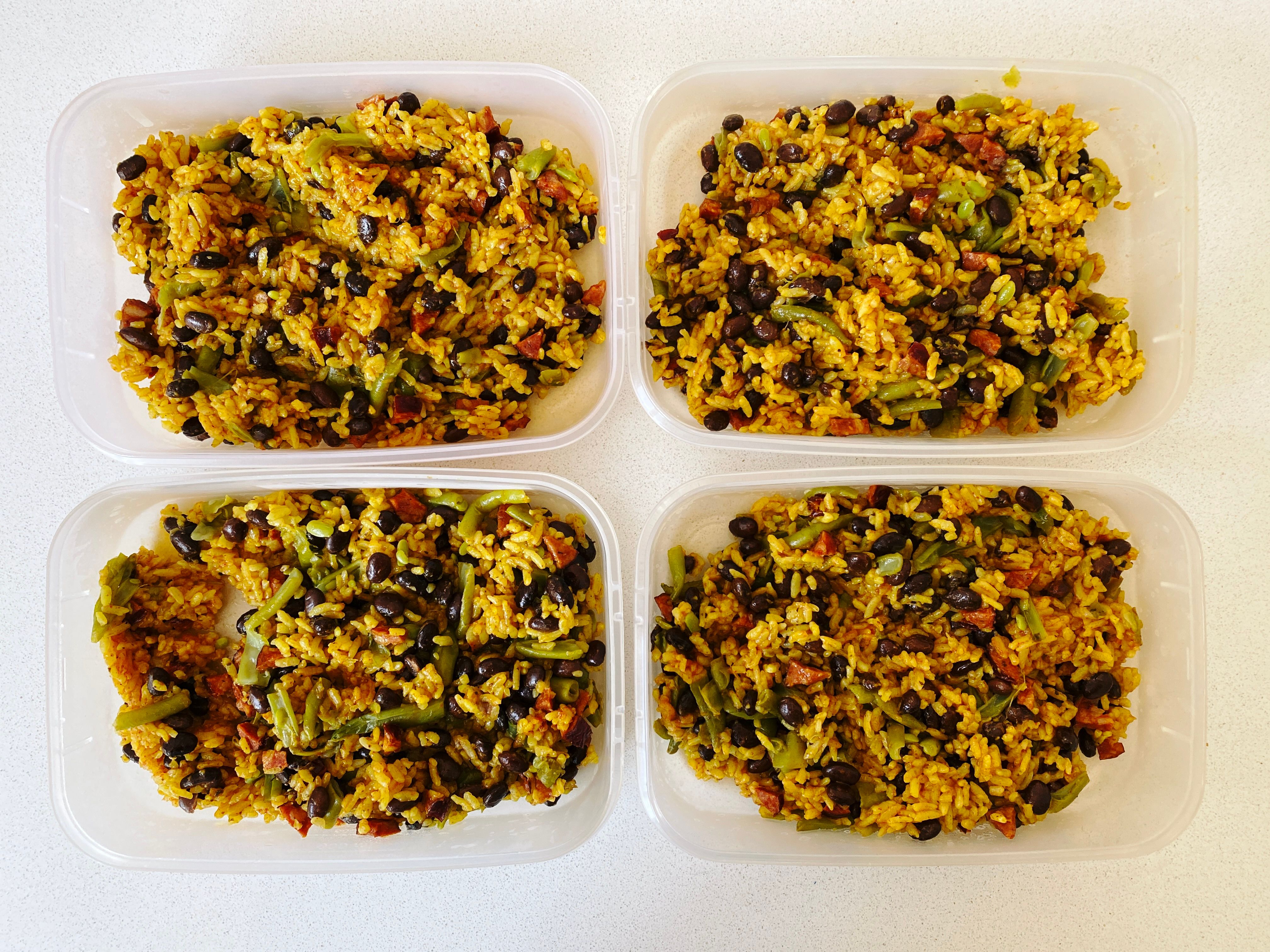 A photo of four plastic food containers with yellow rice, black beans, sliced green beans, and chunks of chorizo.