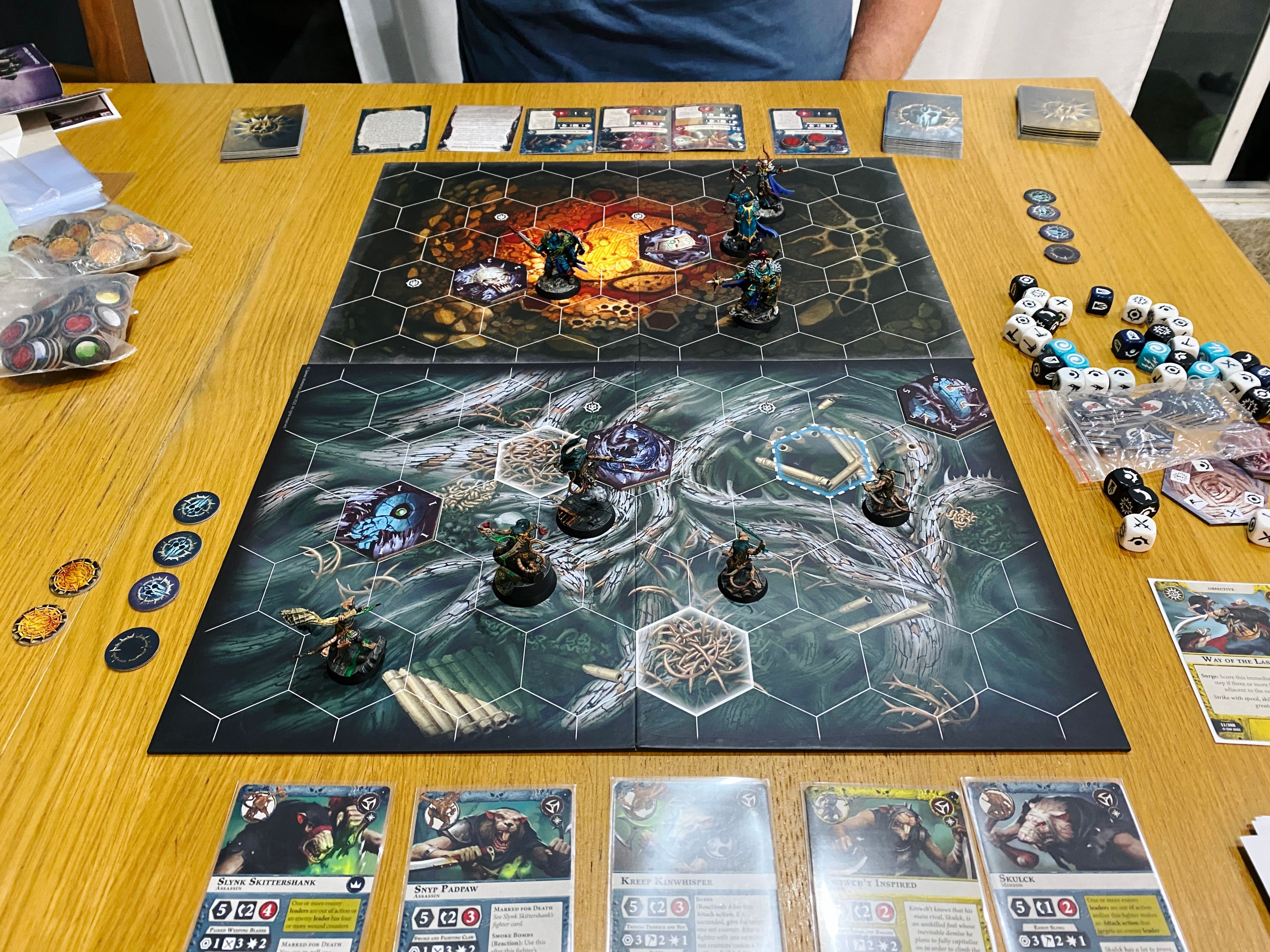 A photo of a Warhammer Underworlds board and miniature. The board is covered in hex tiles and the art on it looks like tree roots and the inside of a cave. Closest to the camera are five Skaven (rat-men assassins) in various dynamic poses (one is leaping over a smoke bomb he's just set off), and facing them are three heavily armoured human warriors of Chaos, and a less heavily-armoured wizard who is wielding a staff and looks like she's in the middle of casting a spell.