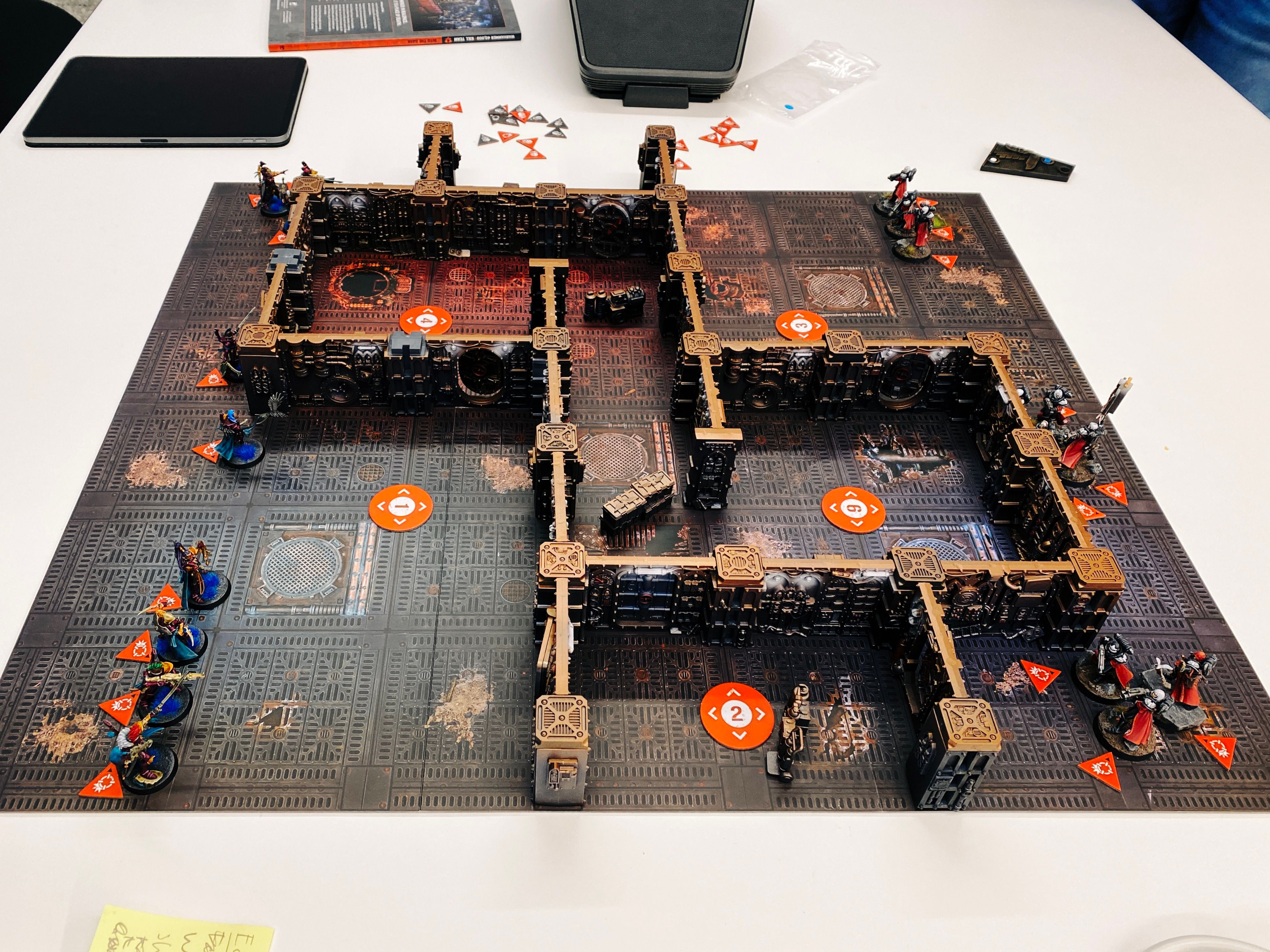 A photo of a game of Warhammer 40,000: Kill Team. The board looks like the metal of the inside of an ancient steampunk starship, and the terrain is a bunch of walls all connected together in the same steampunk style to form number of interconnecting rooms. On the left are nine Aeldari (think space elves) in elegant fuchsia-coloured armour, and at the right are ten heavily-armoured human warrior women in black armour and red cloaks, all with striking white hair.