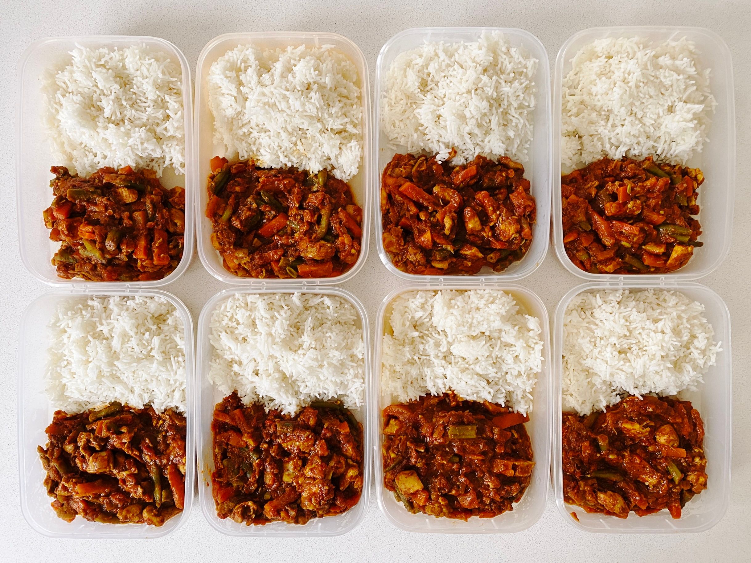A photo of eight Tupperware containers sitting on a bench. They're all filled with half rice and half tomato-based chicken curry with carrots and beans in it.