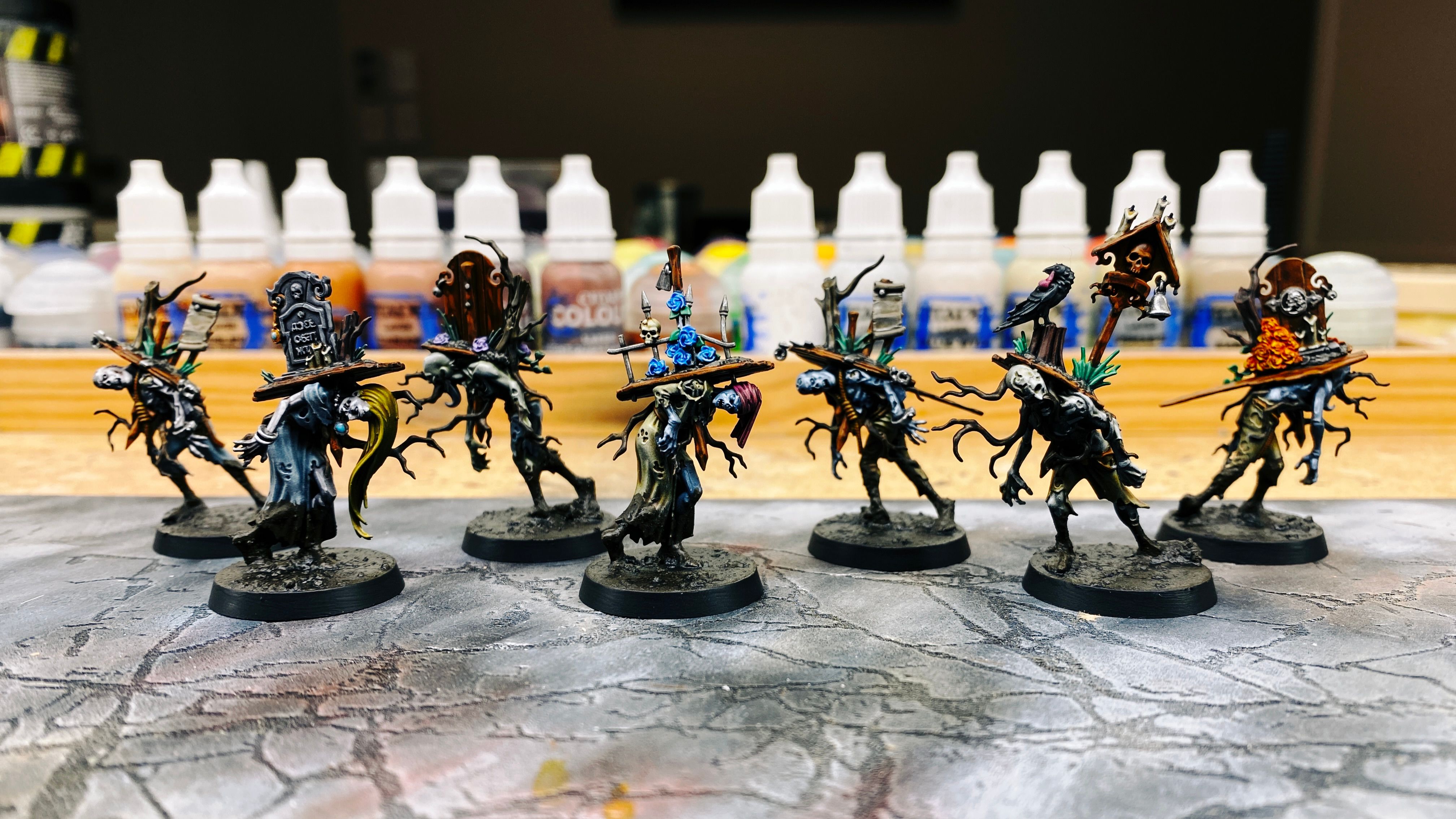 A photo of seven hunched-over rotting zombies. They've got the standard-issue zombie decaying clothes, but all three have part of the lids of the coffins they were in on their backs, which are attached to them by way of stakes driven through the top of the lid and through their chests. They also have tree roots growing through their bodies. The bases are all done up to look like fresh wet mud, which is splattered part-way up their legs so it looks like they've been slogging through it.