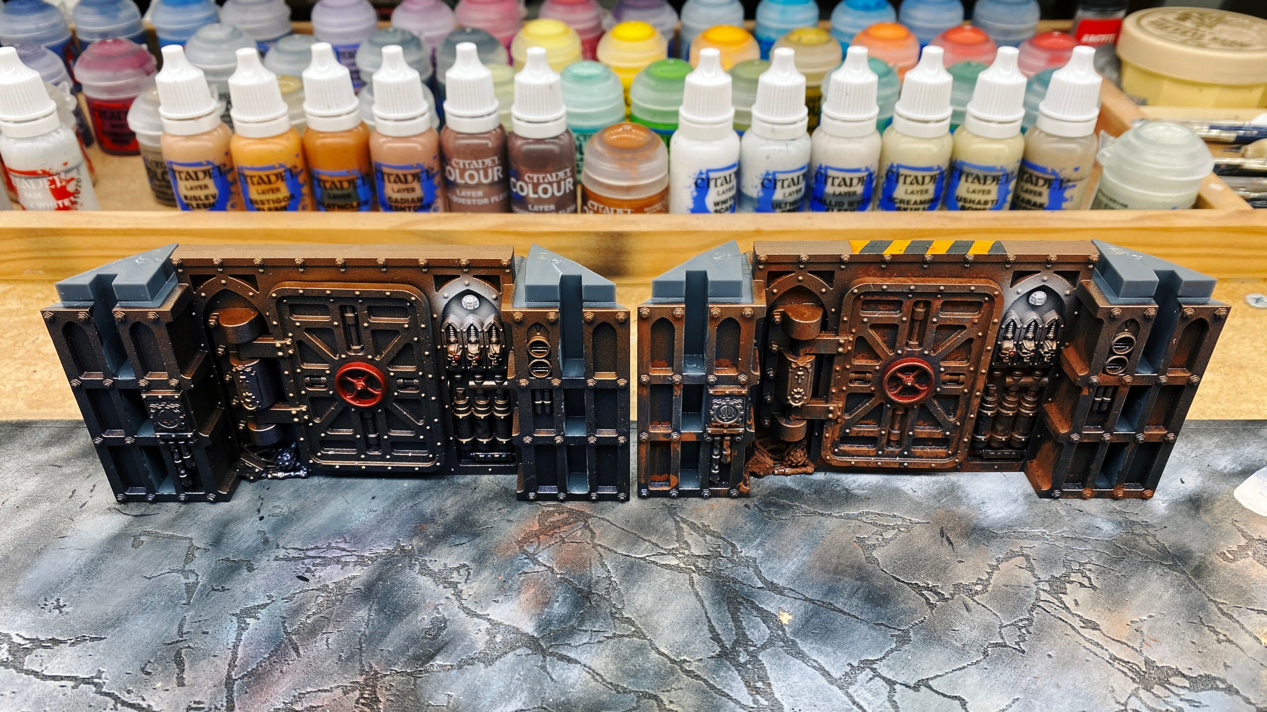 A photo of two of the sections of the wall terrain from Into The Dark. It looks very steampunk with lots of pipes and skulls and both are painted the lower colour being a dark metallic blue and further up the wall it fades into a lighter copper colour. The section on the left looks almost unfinished compared to the one on the right, that looks a lot more "lived in".