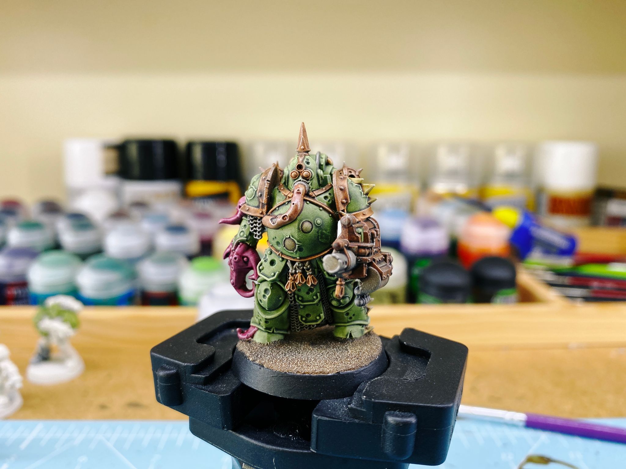 A photo of a Plague Marine from Warhammer 40,000. He's a big green lumbering armoured thing, he's got a grenade launcher-looking gun in one hand, the other is a tentacle, and he's got a fleshy tube thing going into his helmet like it's supplying oxygen (it's totally not oxygen though).