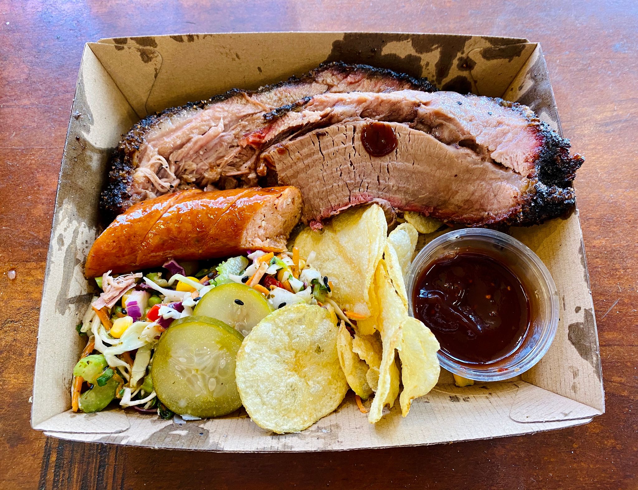 A photo of a cardboard food container with slaw, chips, beef brisket, and some sausage.
